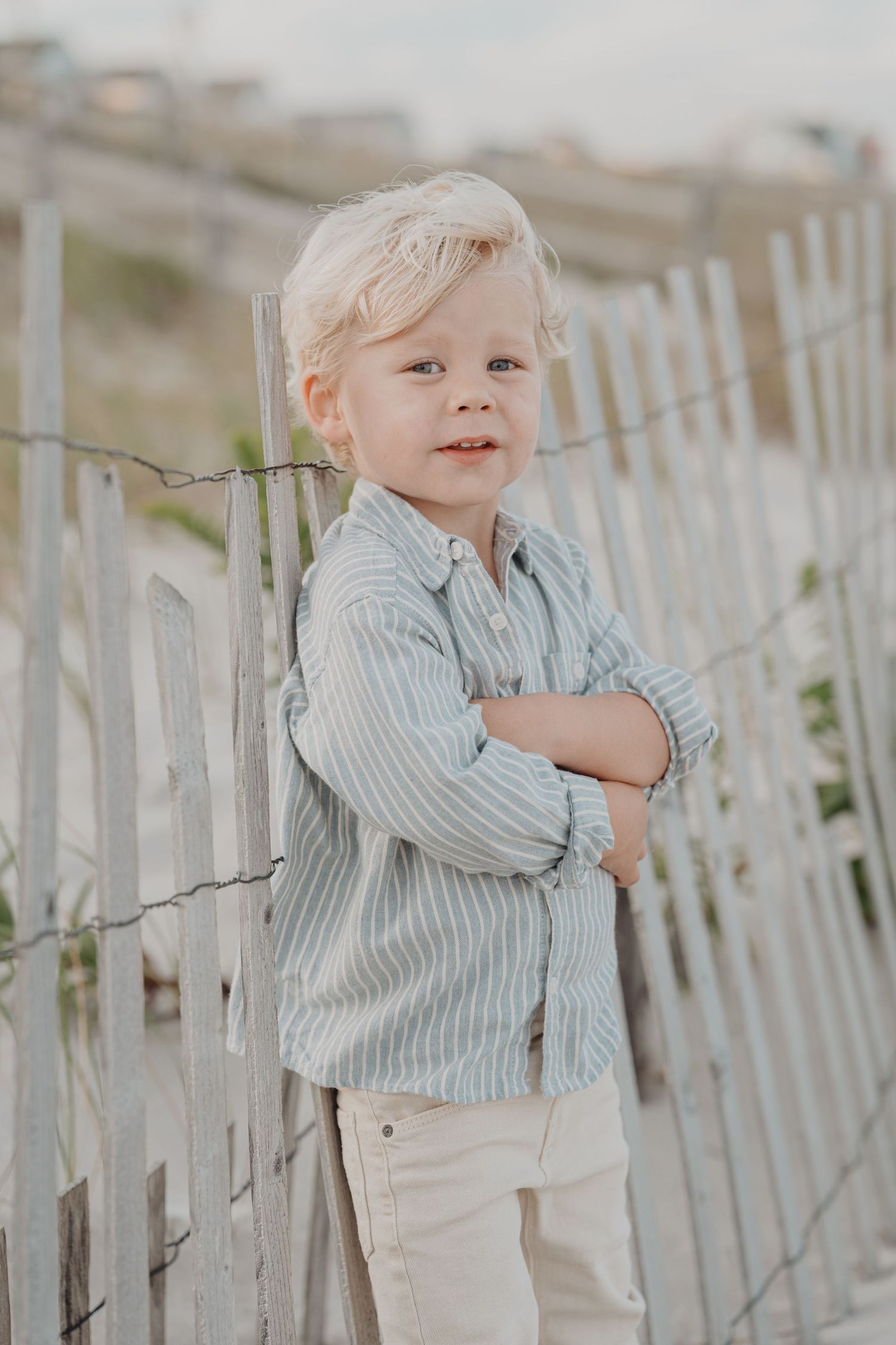 Young child in striped shirt standing by white beach fence during golden hour.