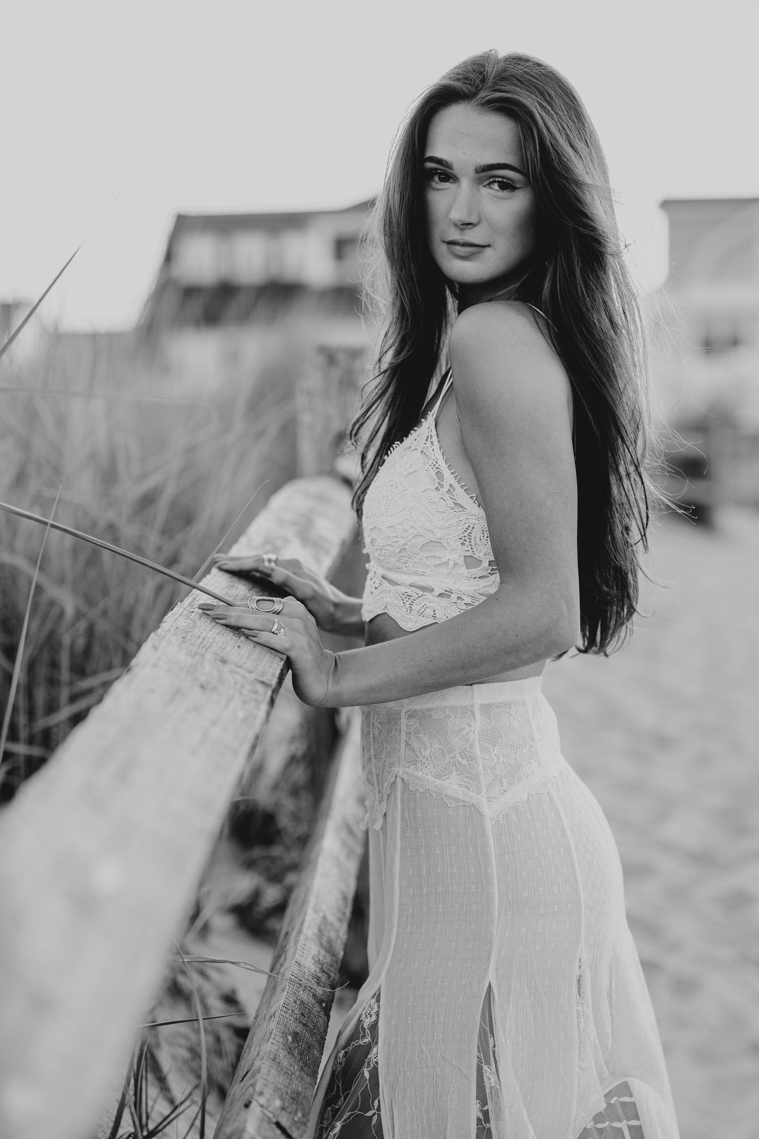 Black and white photo of a brunette woman in a white lace dress leaning on a wooden beach fence.