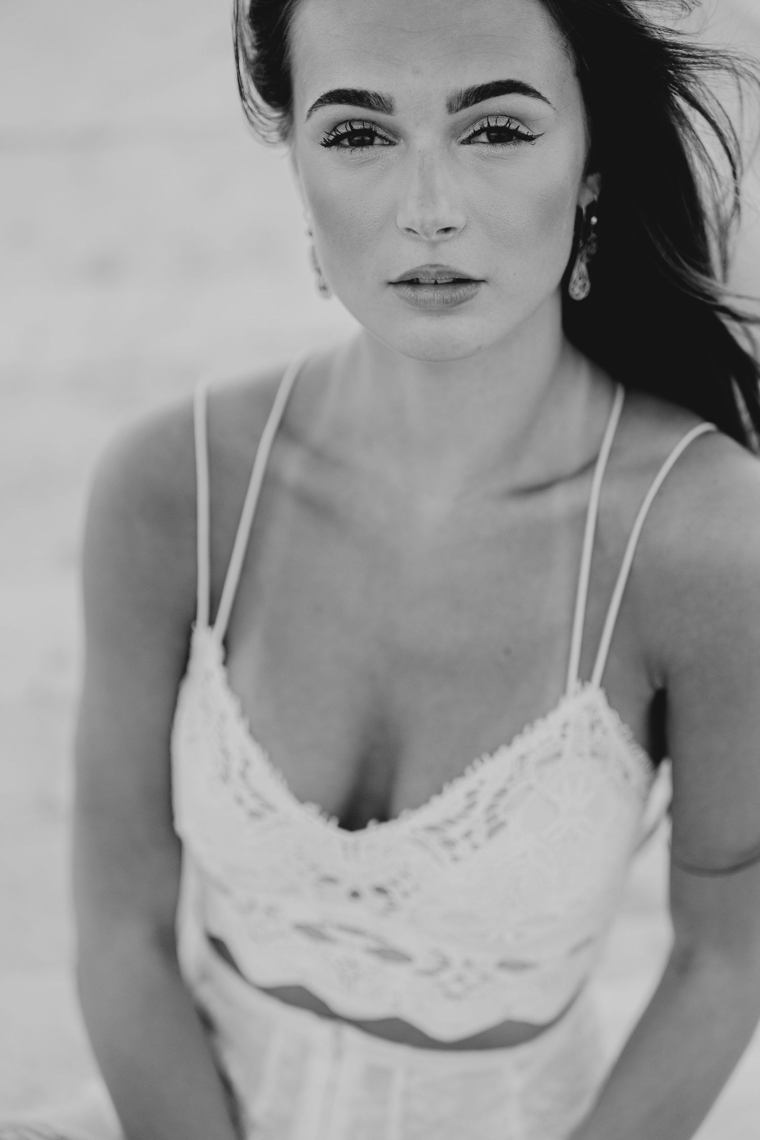 Black and white close-up portrait of a woman in a white lace camisole with elegant earrings on a beach.