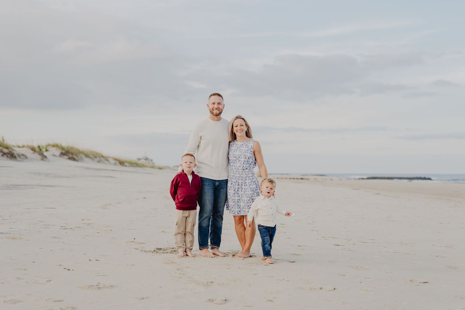 Family of four smiling on a sandy beach, parents holding hands with two young boys on a cloudy day.