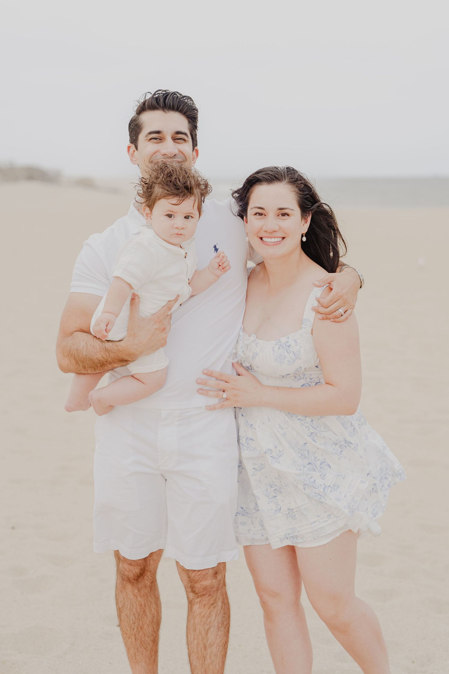 Happy family of three posing on a sandy beach, parents in white outfits holding a baby, smiling outdoors.