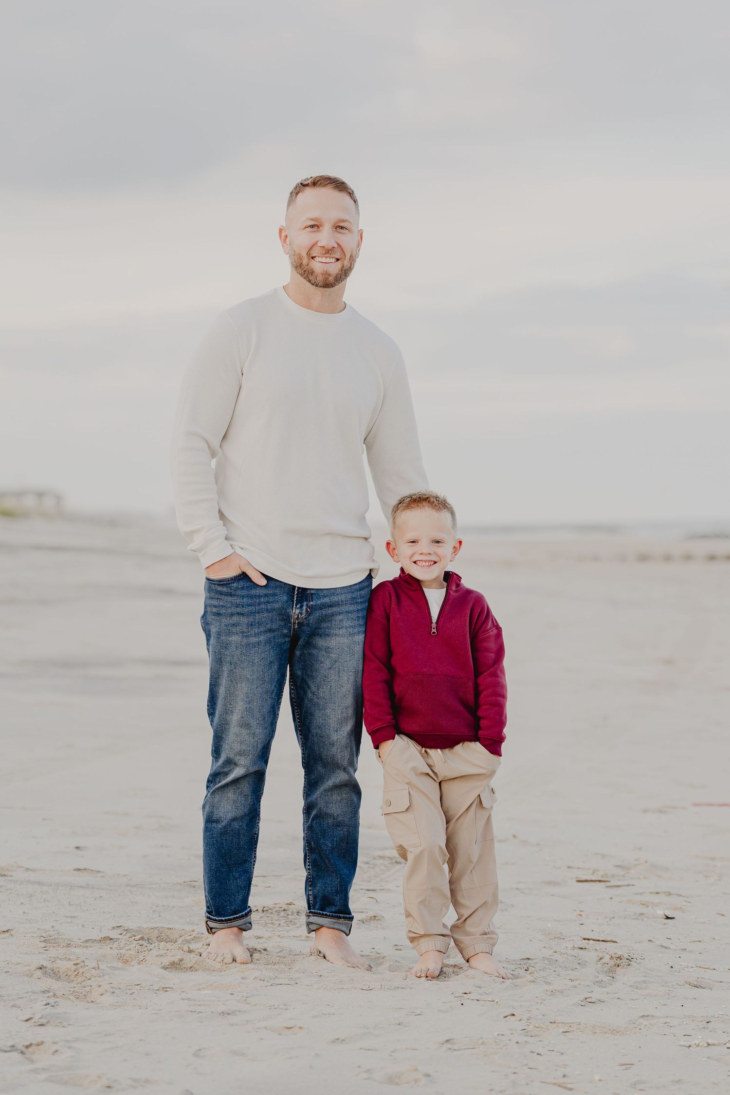 A smiling man and young boy stand barefoot on a sandy beach, wearing casual clothes in a heartwarming father-son portrait.