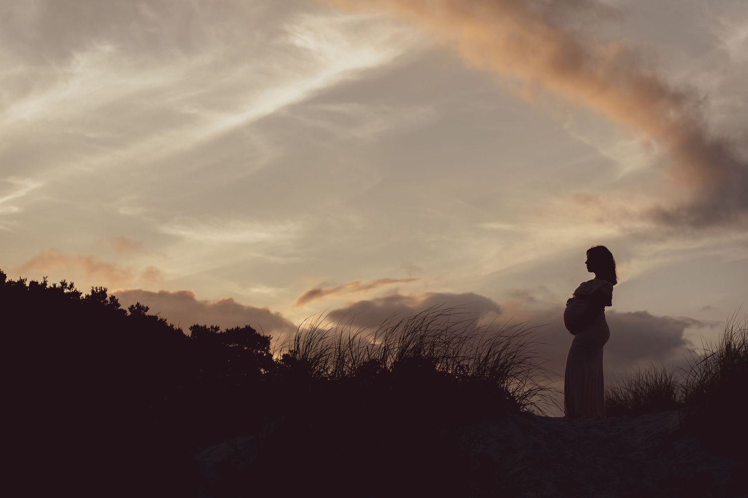 Silhouette of pregnant figure standing against dramatic sunset sky with mountains.