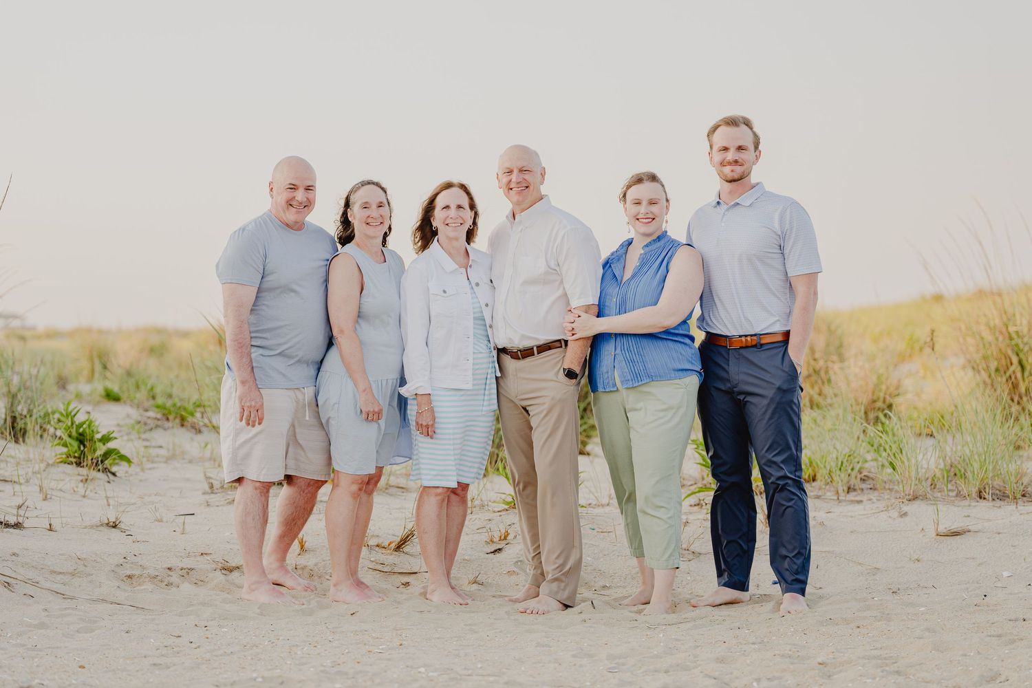 Six family members smiling together on a sandy beach during golden hour in casual summer attire.