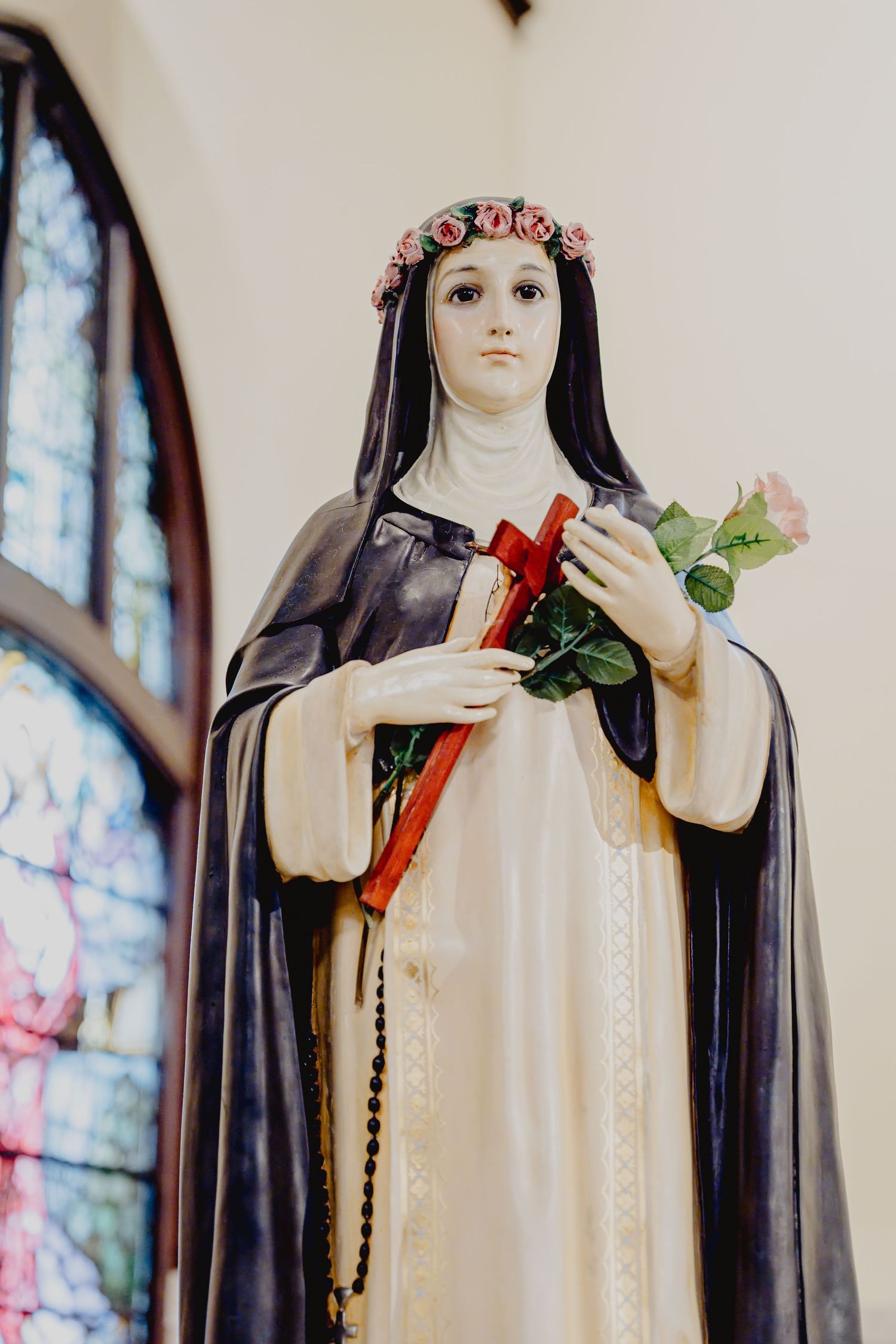 Colorful statue of Saint Therese of Lisieux holding a cross and roses beside a stained glass window.