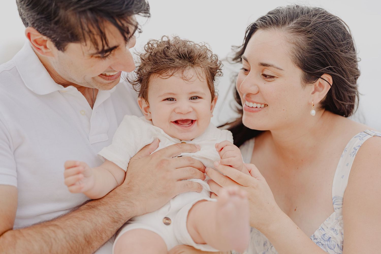 Happy family portrait with smiling baby held by loving parents in a bright, cheerful outdoor setting.