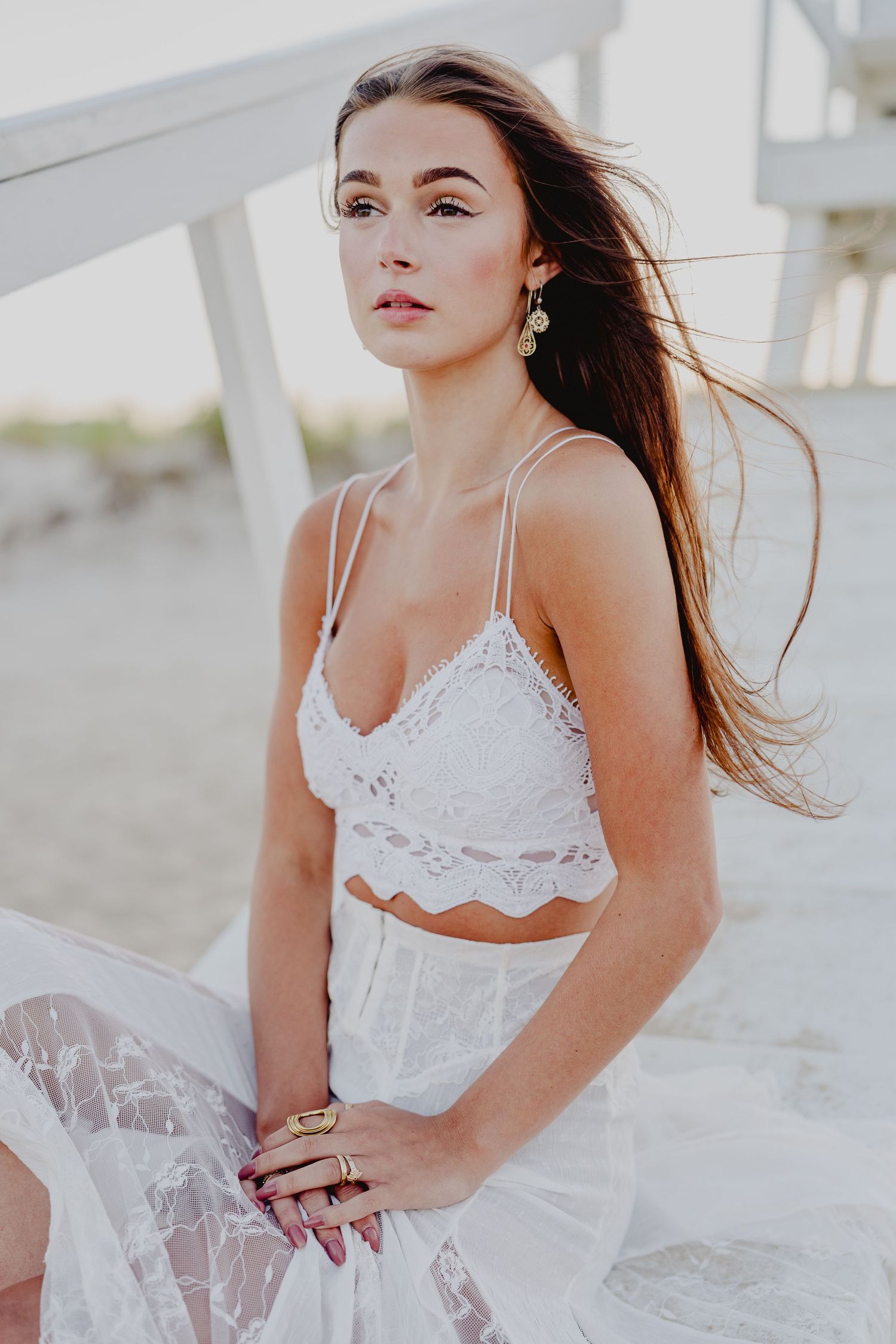 Young woman in white lace crop top and skirt poses gracefully on a beach with wind-blown hair.