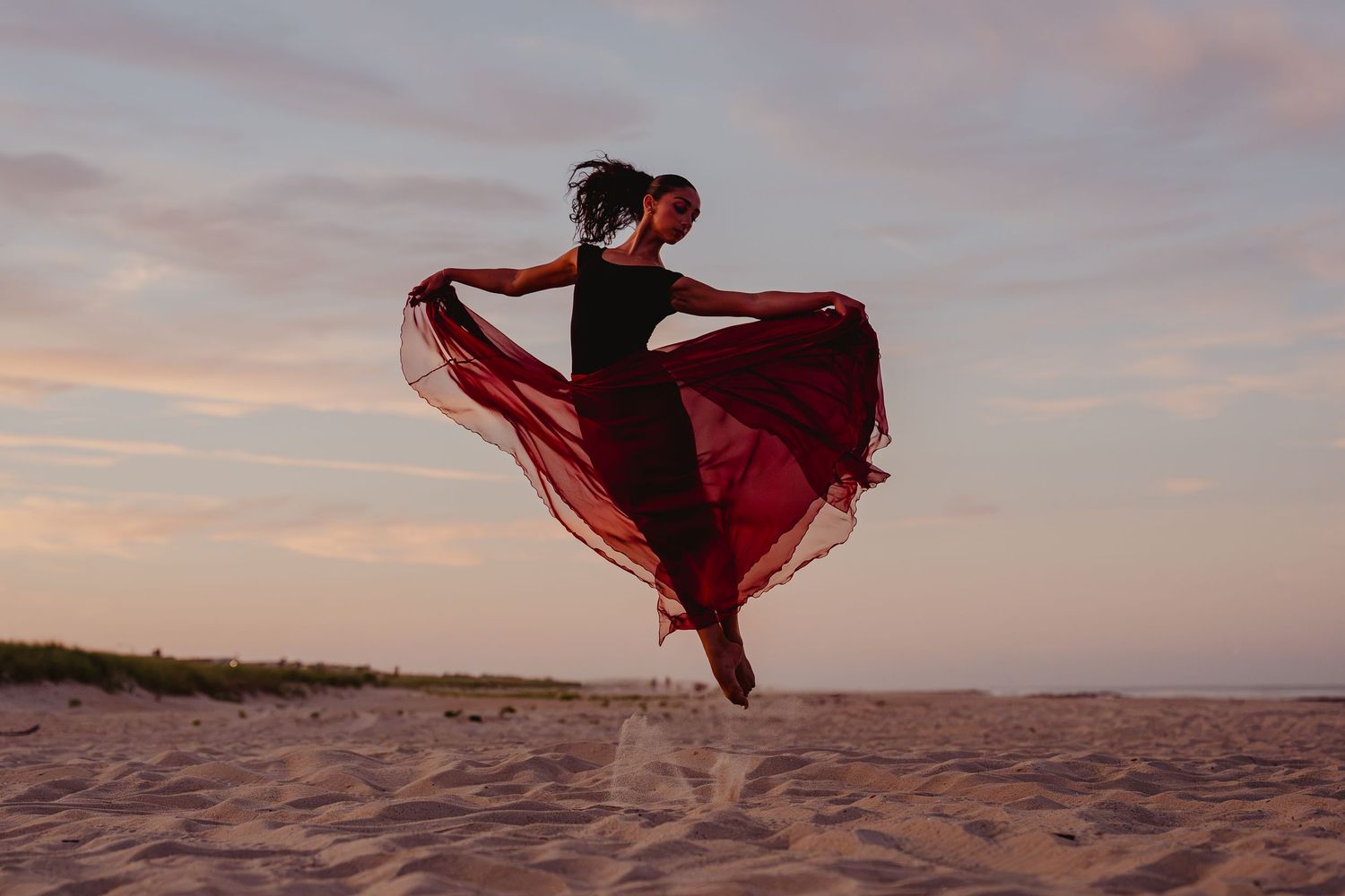 A woman in a black top and flowing red skirt leaps gracefully on a sandy beach at sunset.