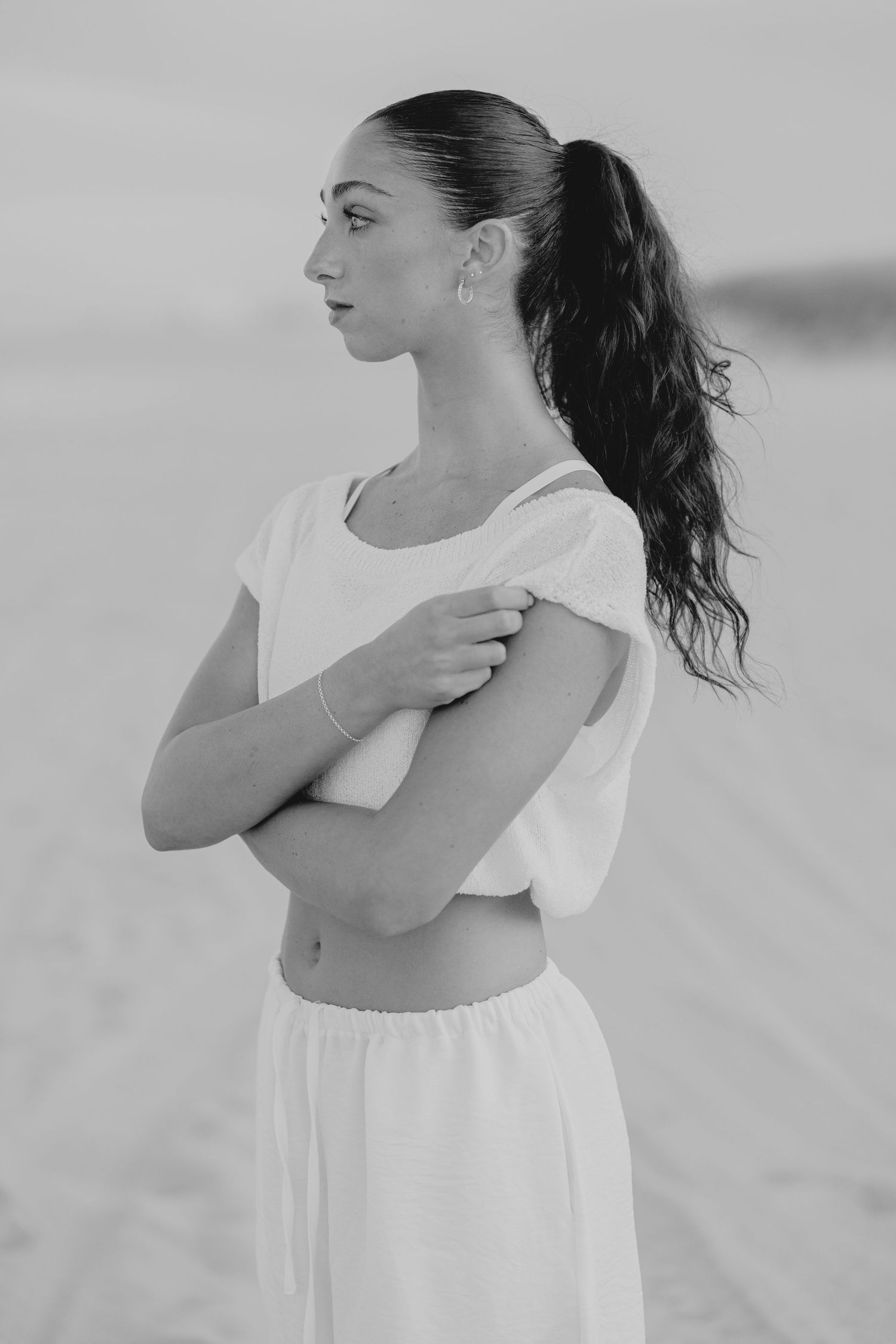Black and white portrait of young woman in white outfit with ponytail on beach.