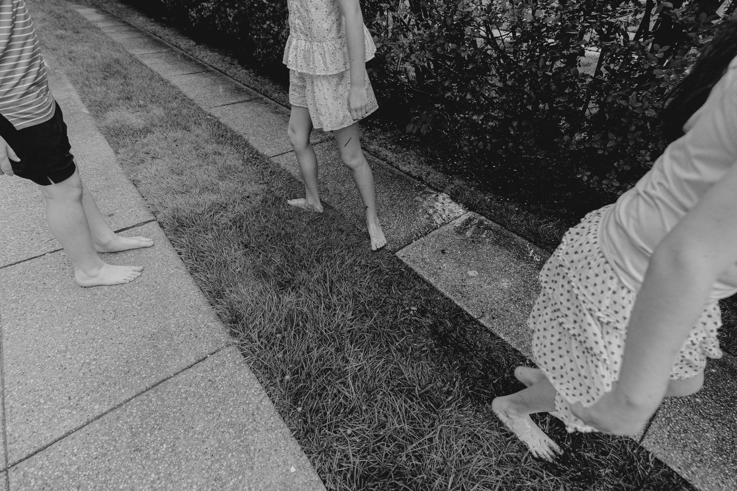 Black and white photo of children walking barefoot on grass beside a sidewalk.