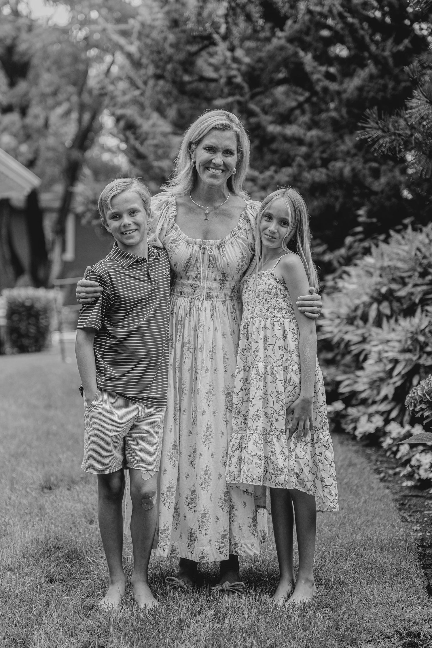 Black and white photo of a smiling woman in floral dress posing outdoors with two children on a lush green lawn.