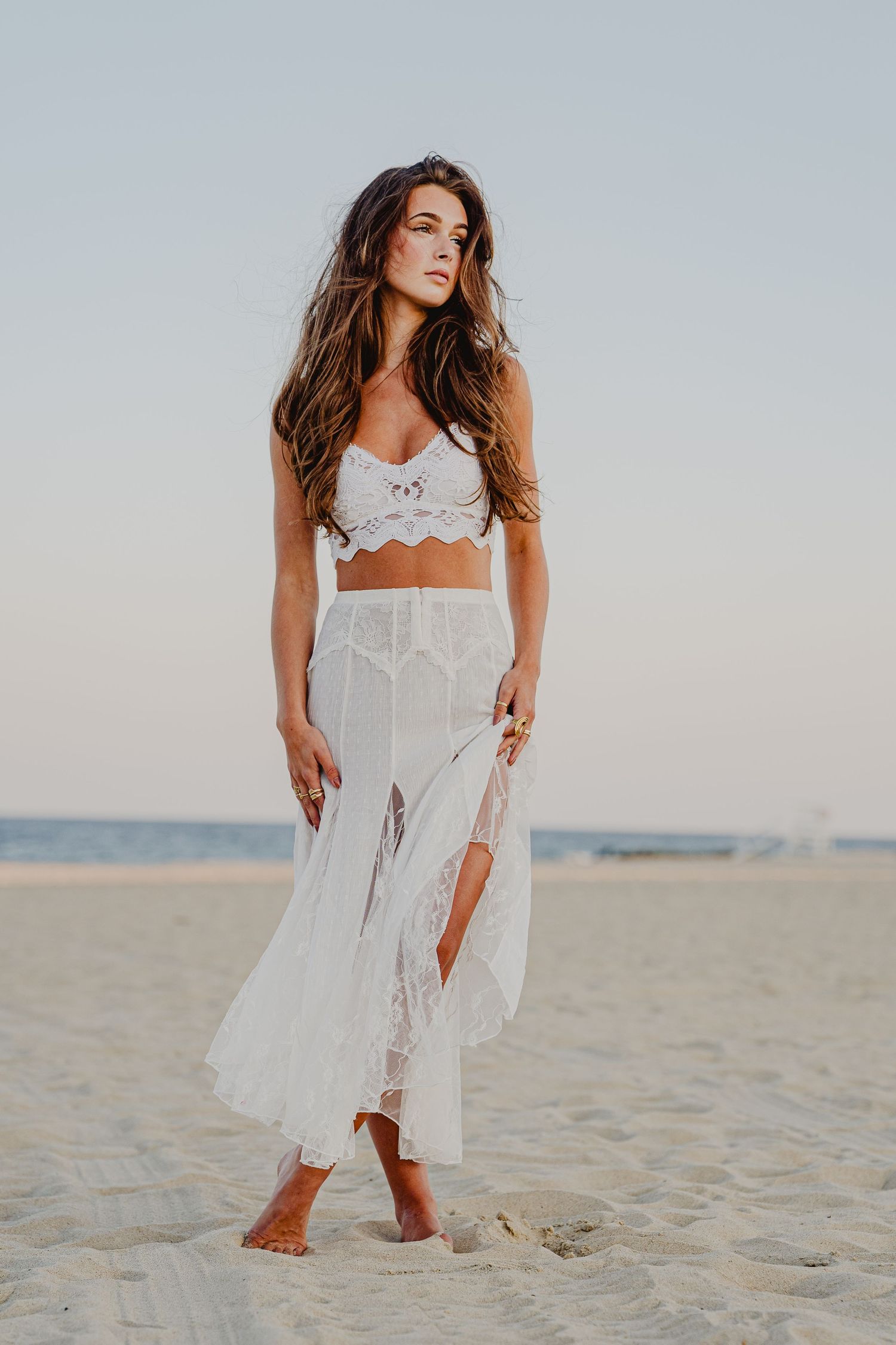 Woman in white lace crop top and sheer maxi skirt standing barefoot on sandy beach at sunset.