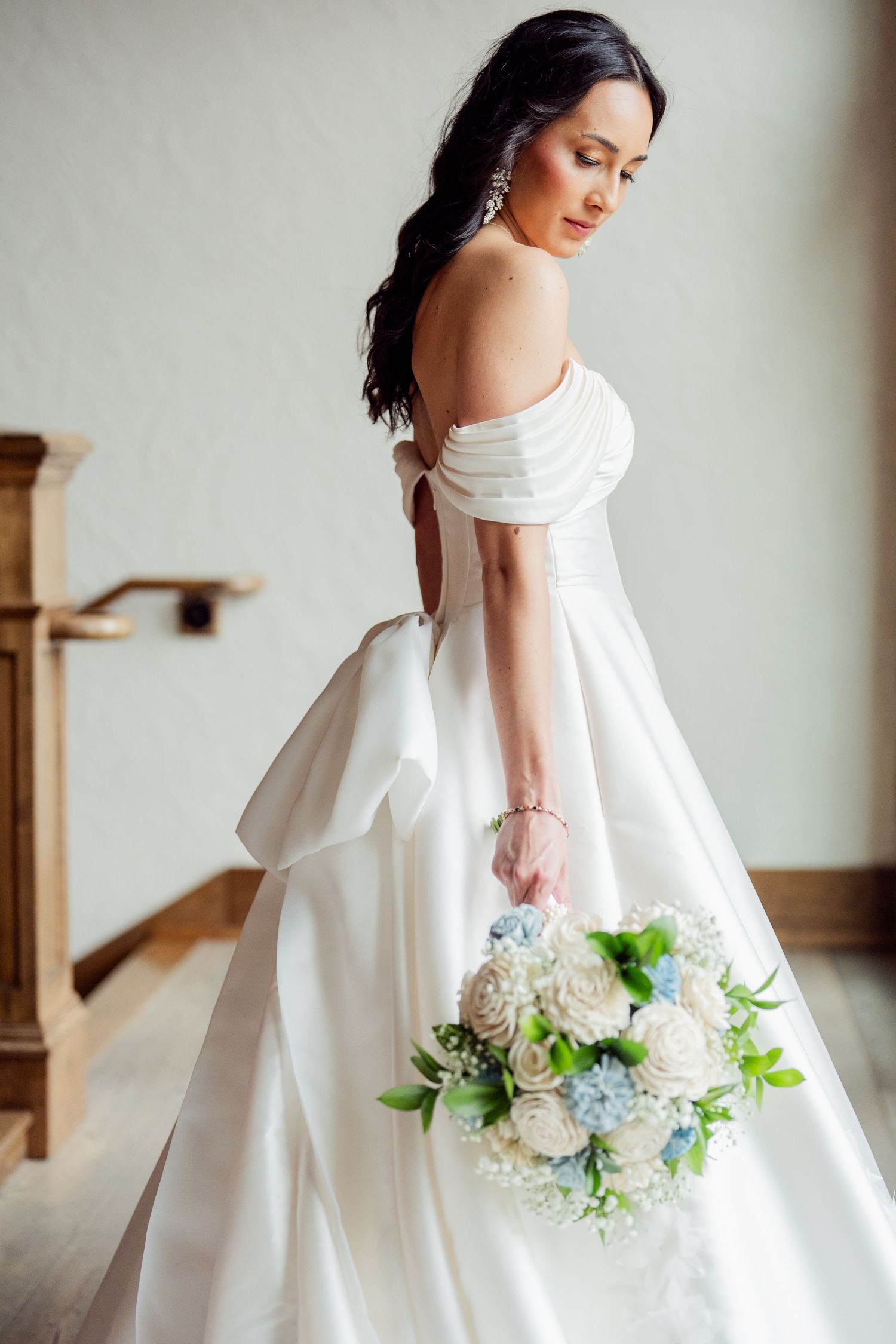 Elegant off-shoulder wedding dress with flowing skirt and blue and white bouquet.