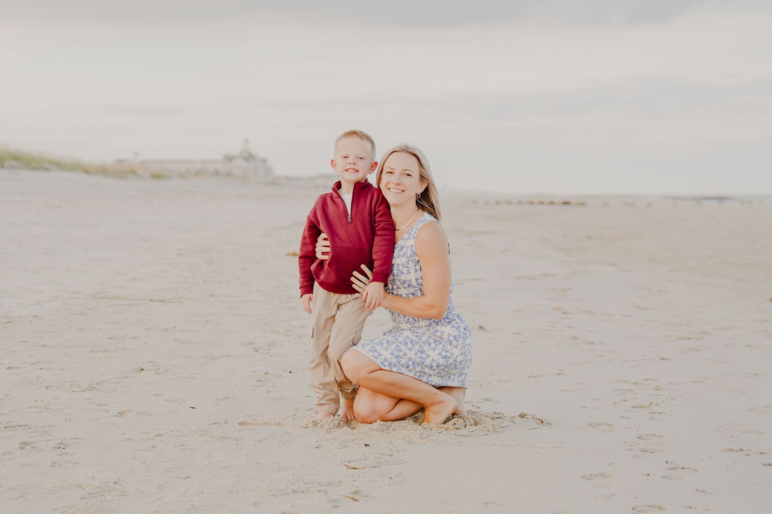 A smiling woman kneels beside a young boy in a red jacket on a sandy beach, posing for a family portrait.