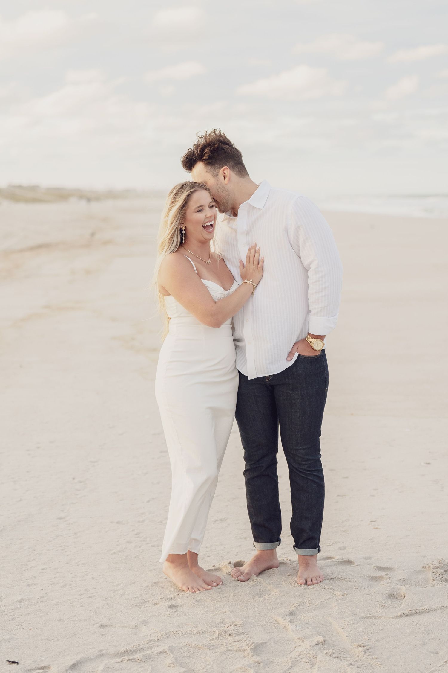 A couple playfully interacts on a sandy beach wearing matching white outfits.