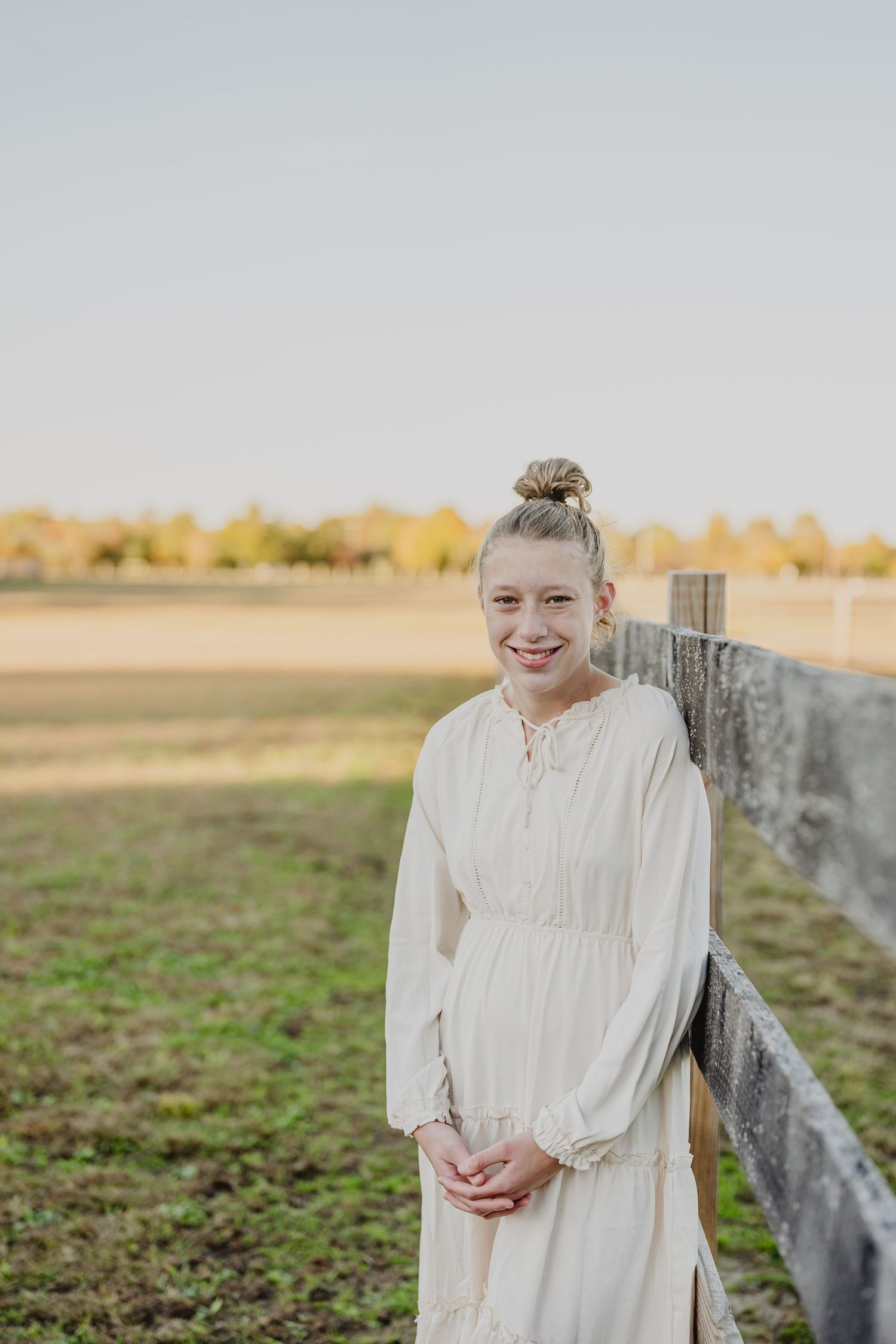 A person in a white vintage-style dress stands against a rustic fence at golden hour in a rural field setting.