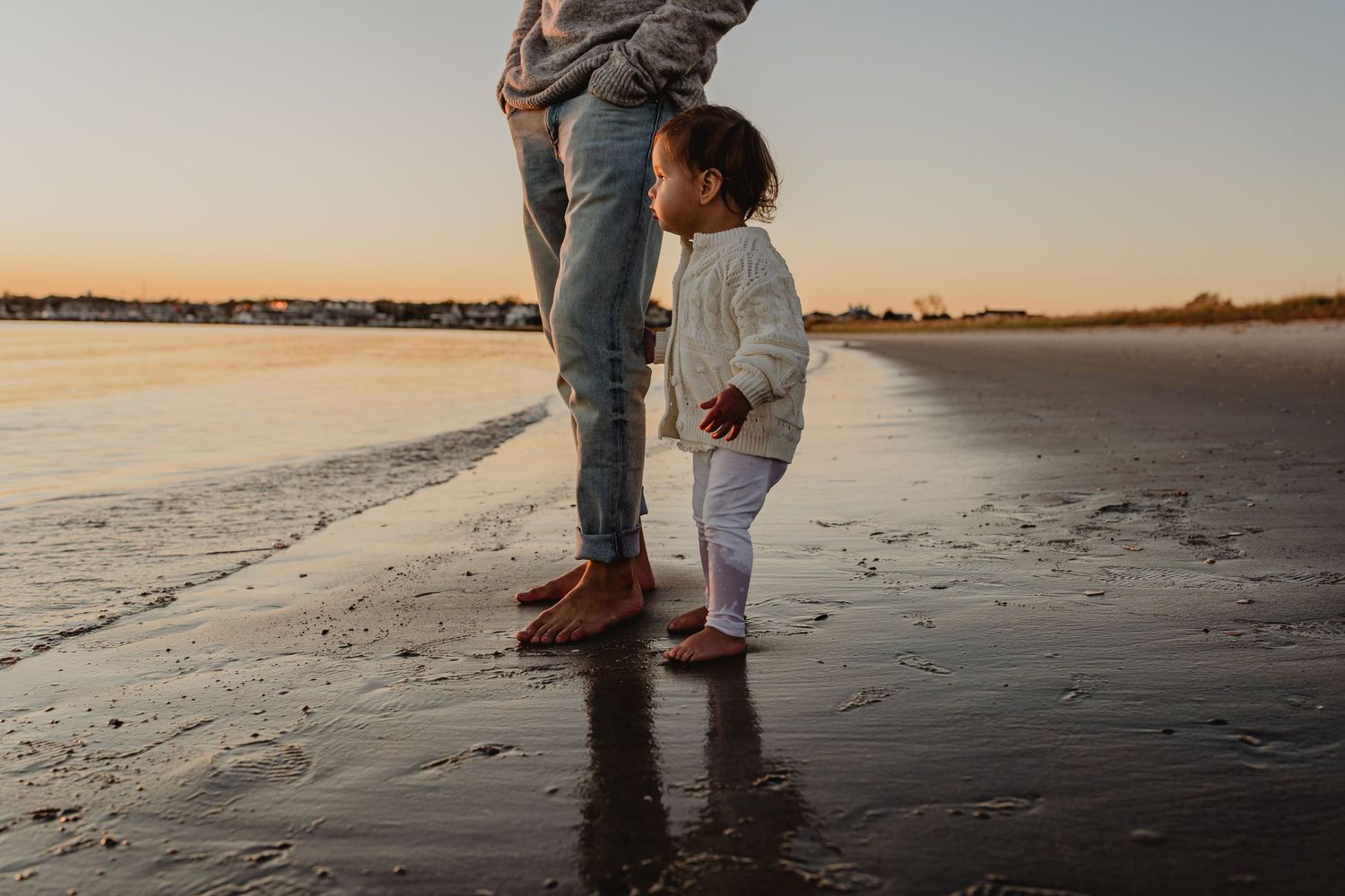 Silhouettes of parent and child walking together on a sandy beach at sunset with reflective water and warm light.