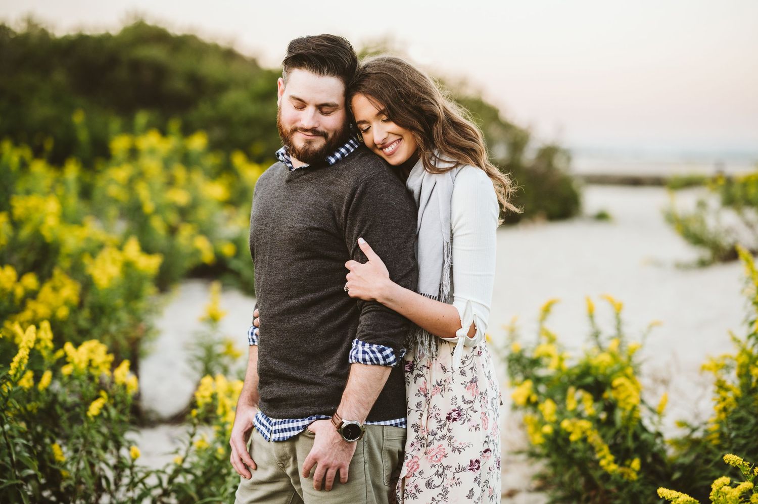 Romantic couple embraces among vibrant yellow wildflowers at sunset by the water.