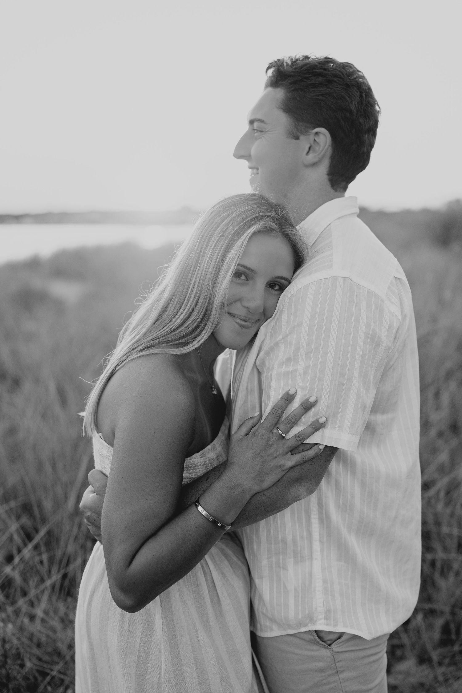Black and white photo of a couple embracing outdoors, woman smiling at camera while man gazes away, near a coastal grassy field.