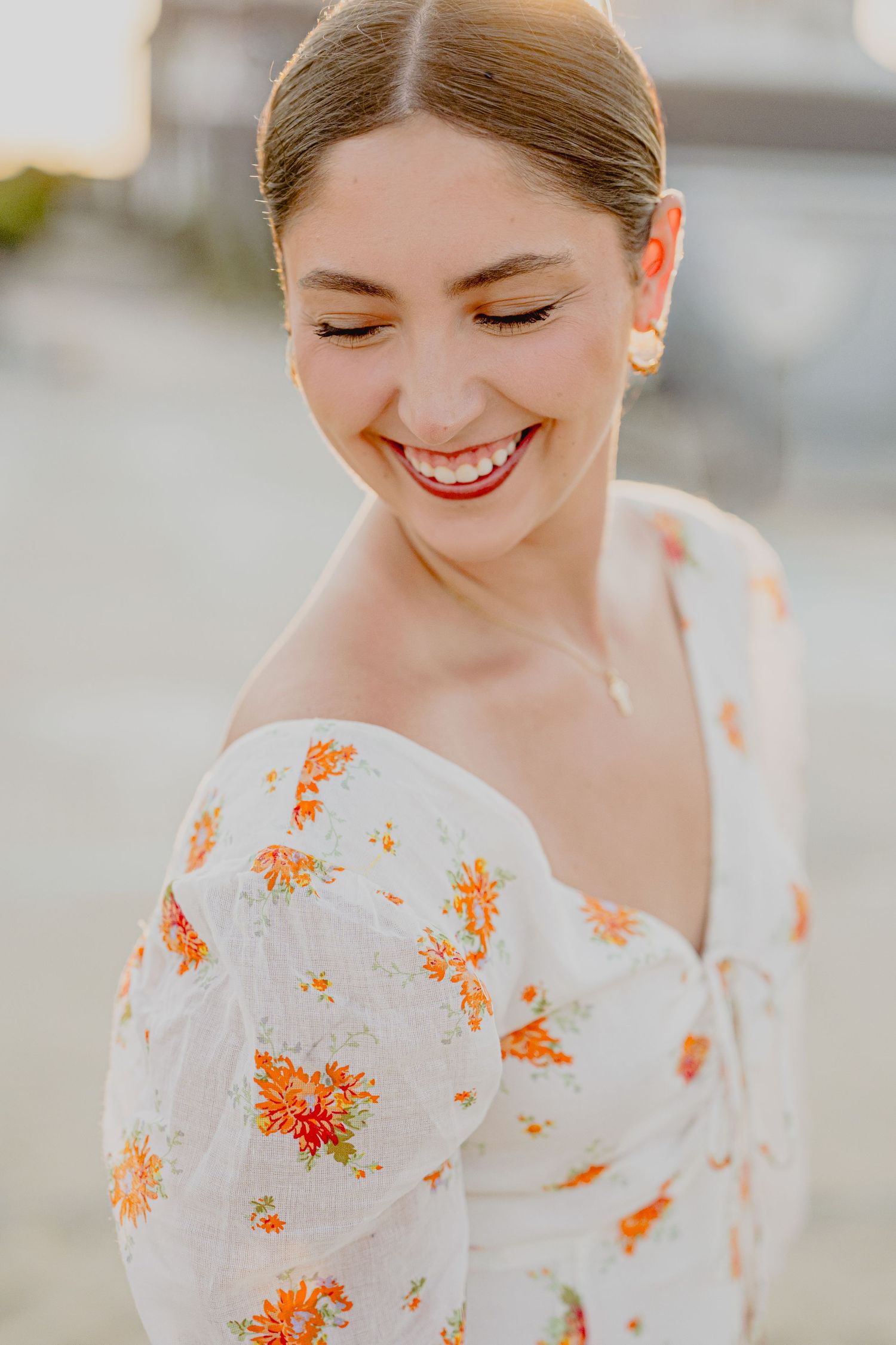 Smiling young woman in a white floral off-shoulder dress with orange flowers, laughing outdoors in warm golden light.