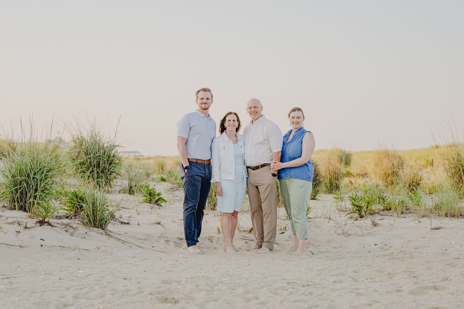 Four adults smiling together on a sandy beach at sunset with dune grass in the background.
