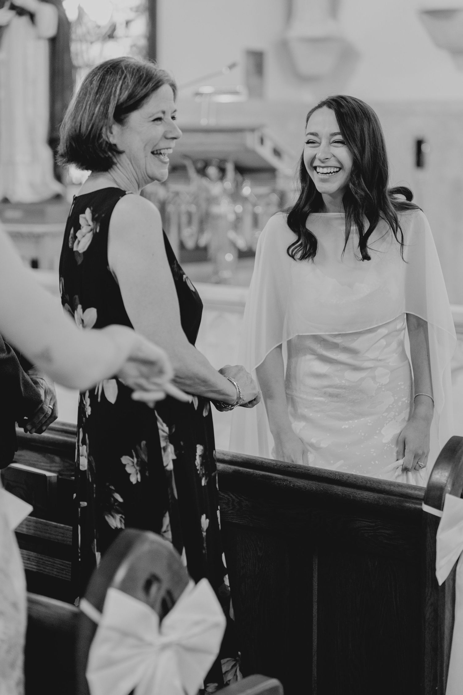 Bride in white gown laughing with guest while holding hands inside a church during wedding ceremony.