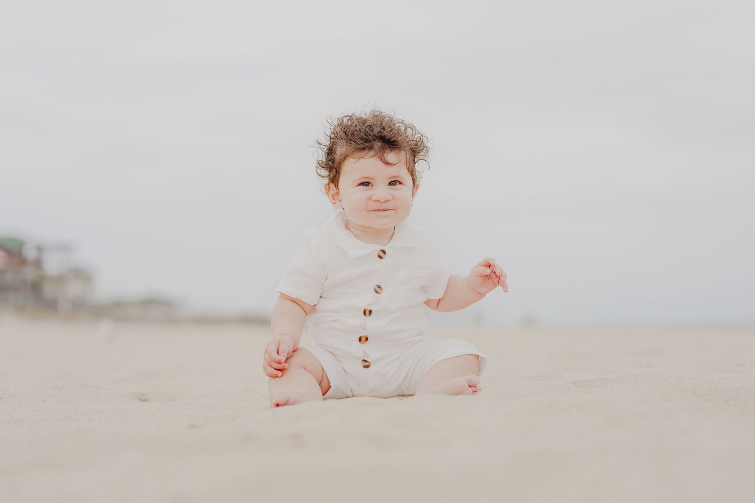 Curly-haired toddler in white romper sitting on sandy beach, smiling softly with overcast sky behind.