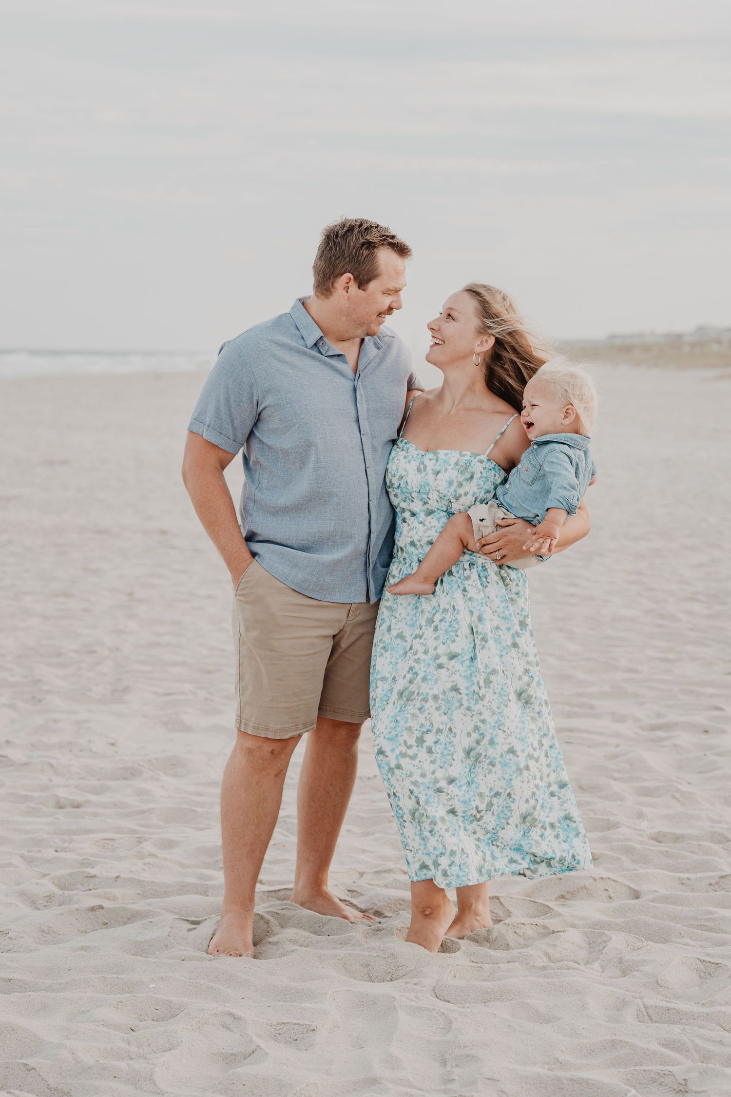 Family of three sharing tender moments on sandy beach in coordinated blue outfits.