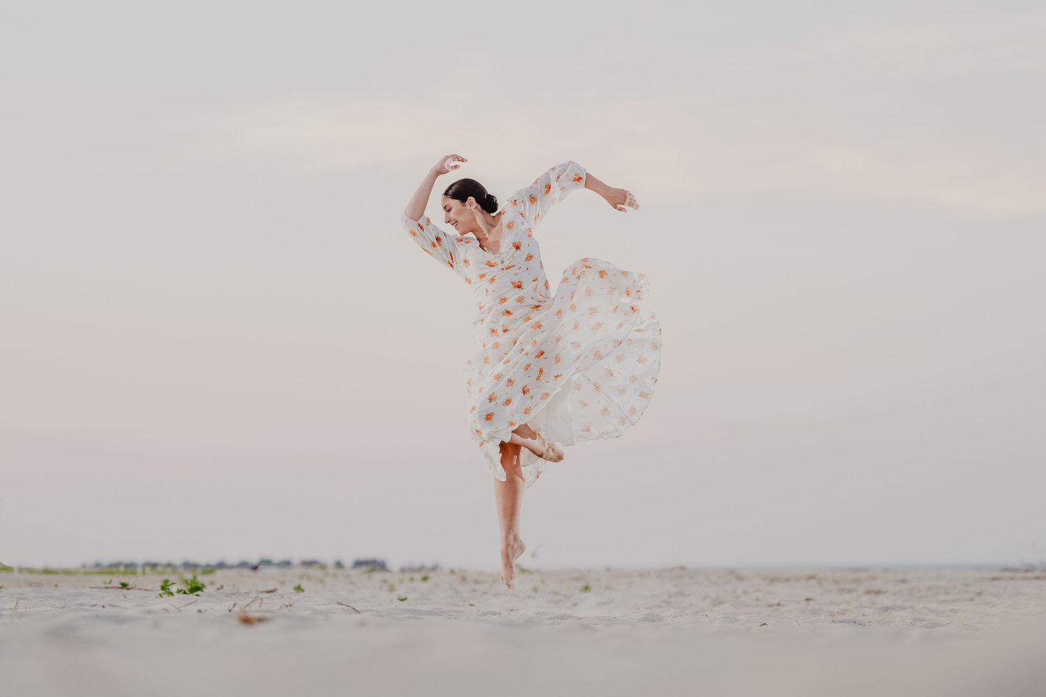 Woman in floral dress dancing gracefully on a sandy beach, arms raised, one leg lifted, against a soft pastel sky.