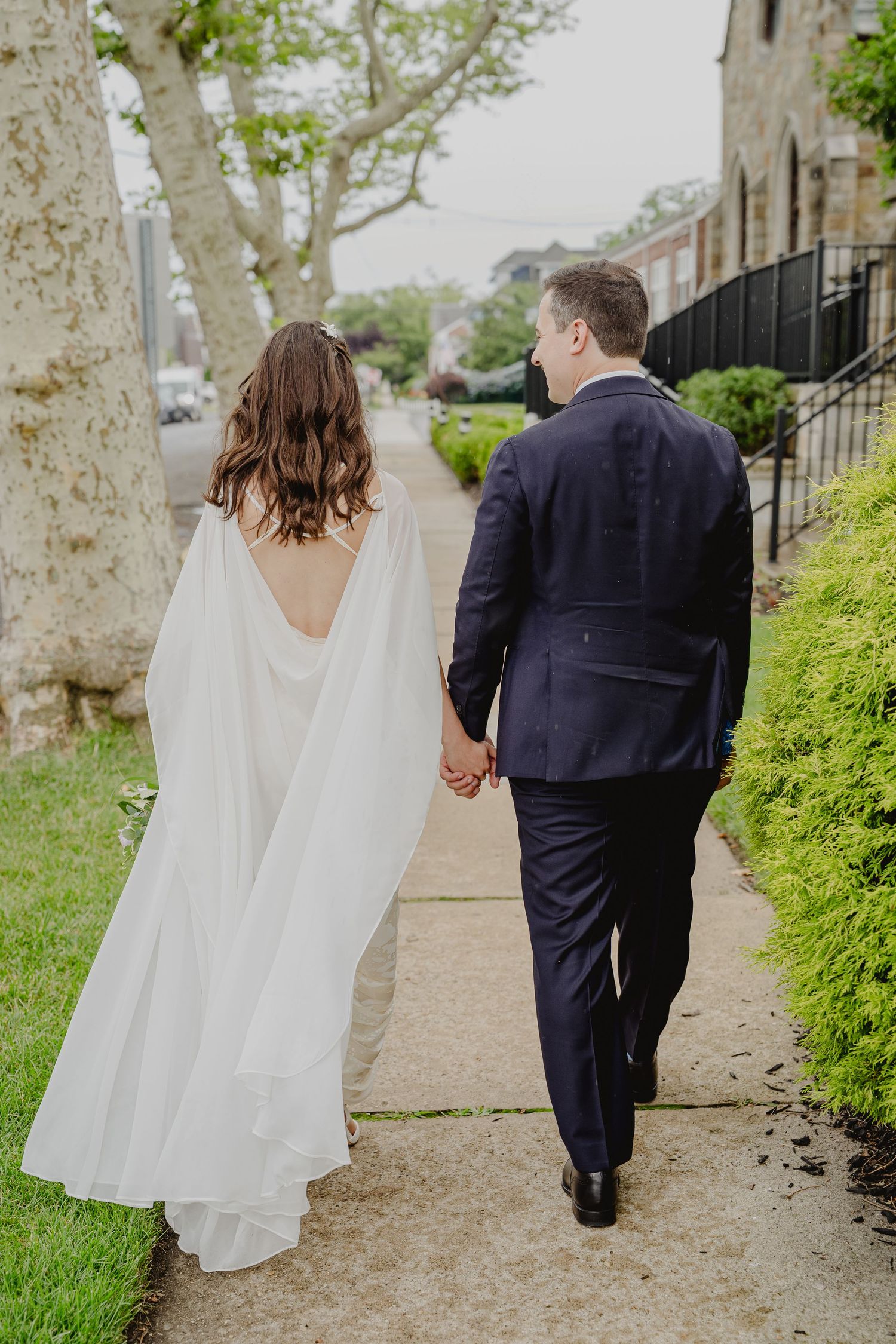 Bride in white cape gown and groom in navy suit holding hands, walking on a sidewalk.