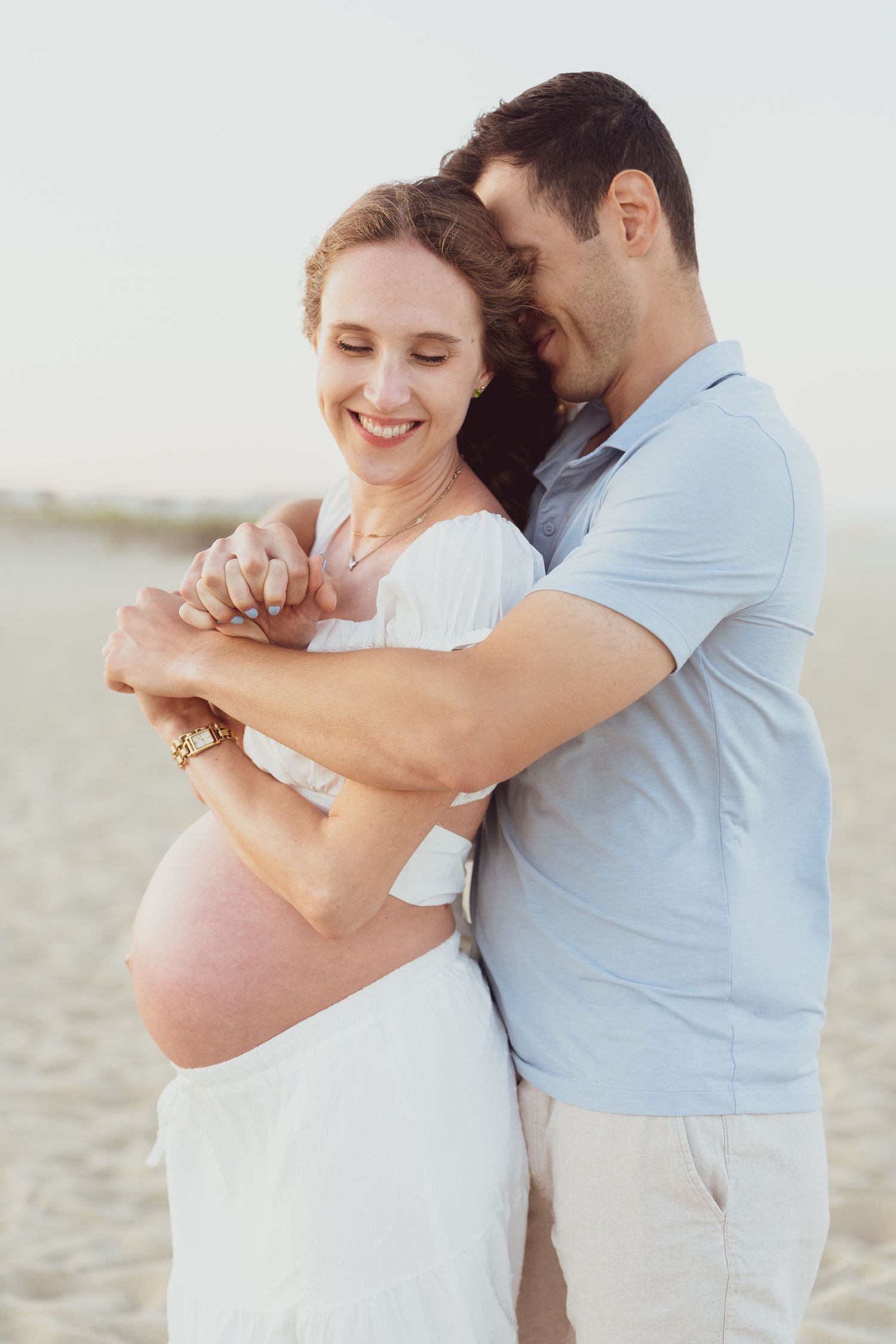 A couple embraces tenderly on a bright beach wearing white and light blue outfits.