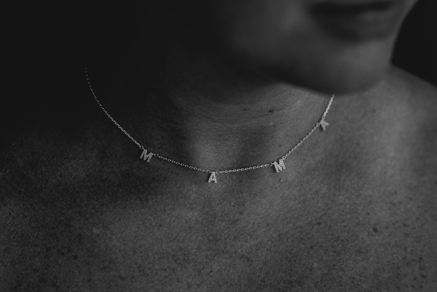 Close-up black and white photo of a delicate necklace spelling out MAMA against skin.