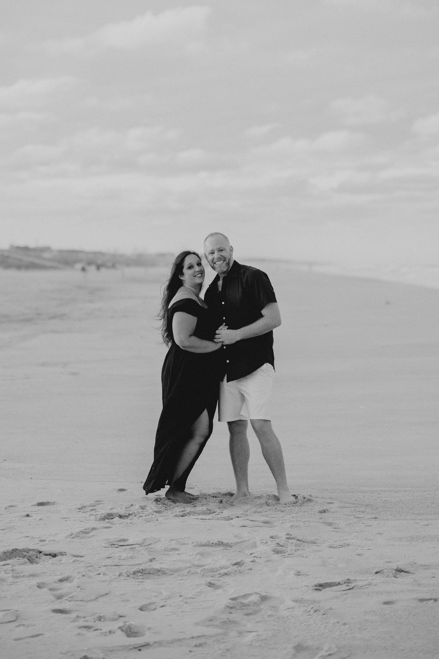 A couple in black attire poses intimately together on coastal sand dunes.