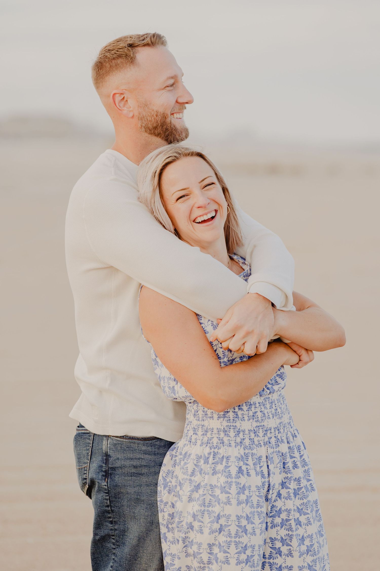 A joyful couple embraces and laughs together on a sandy beach at golden hour.