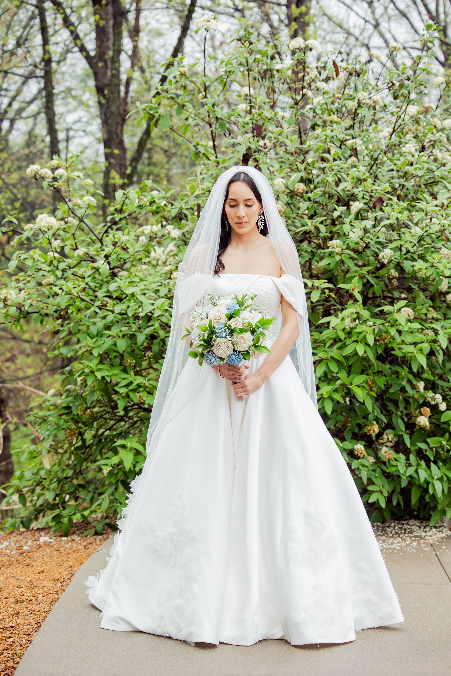 Bride in flowing white dress and veil poses elegantly on garden pathway.