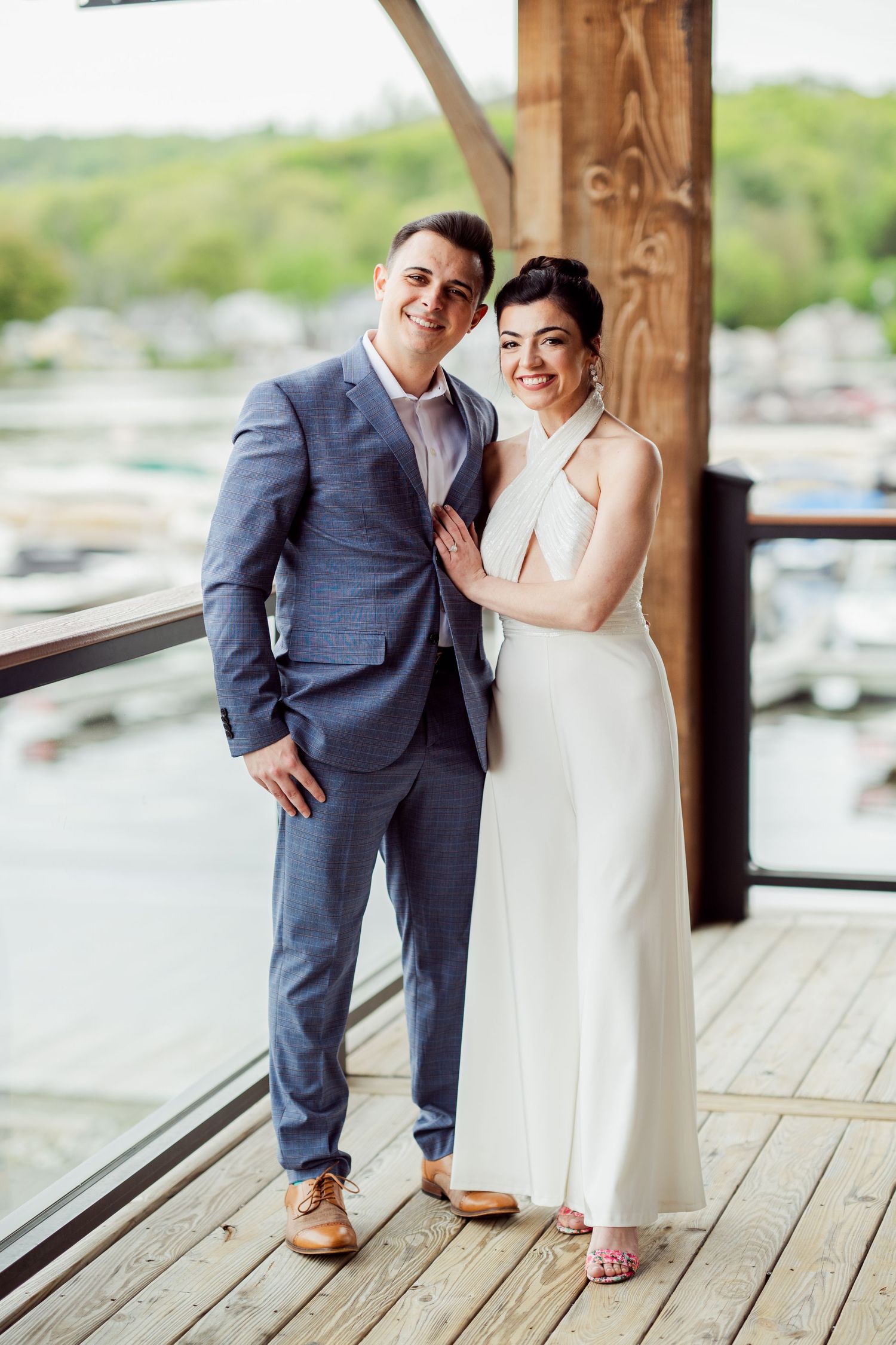A couple poses together on a wooden deck overlooking a marina and lush landscape.