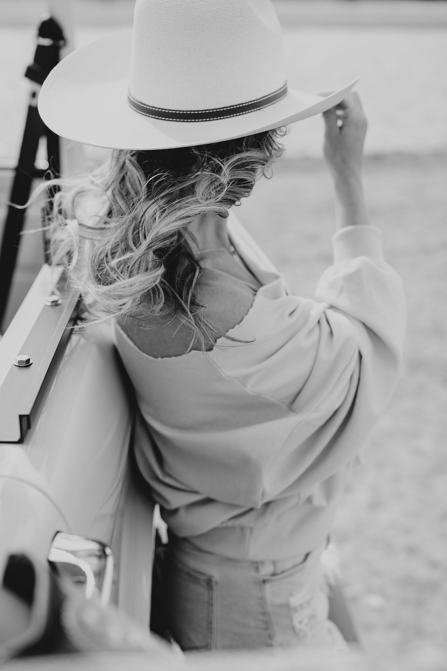 Black and white photo of a blonde woman in a wide-brim hat and loose blouse leaning against a vintage vehicle.