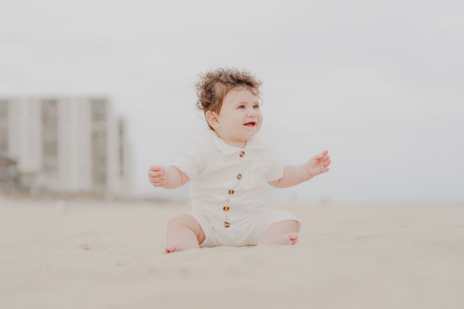 A happy baby in a white romper sits on a sandy beach with arms outstretched, smiling joyfully outdoors.