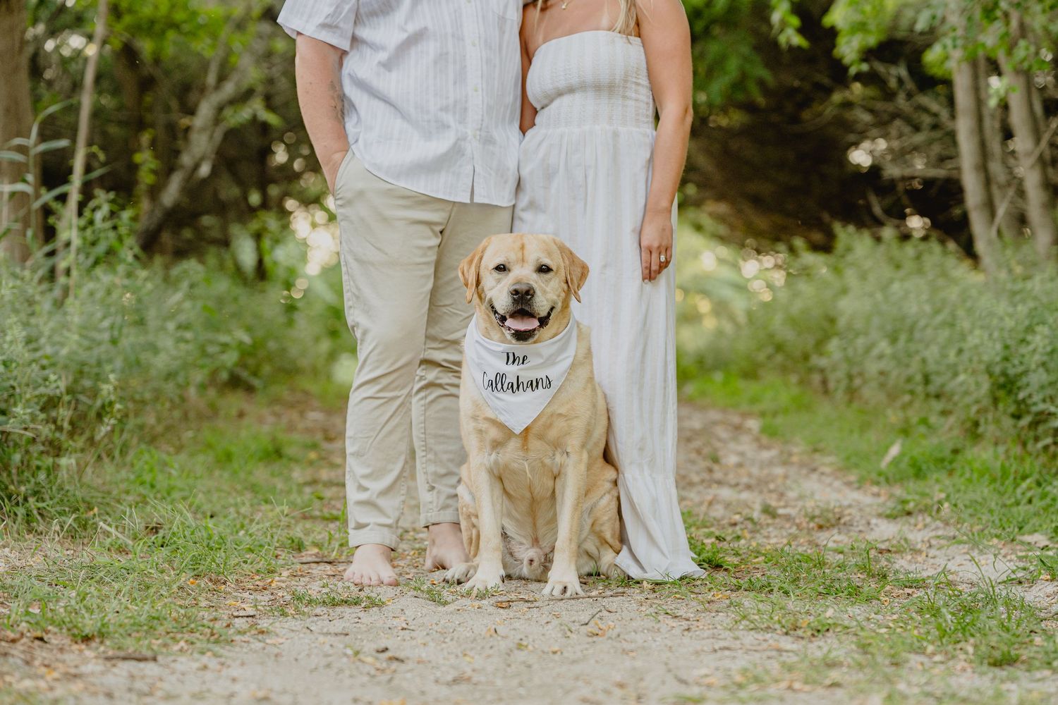 Couple with golden Labrador wearing wedding bandana sits on dirt path surrounded by lush green trees.