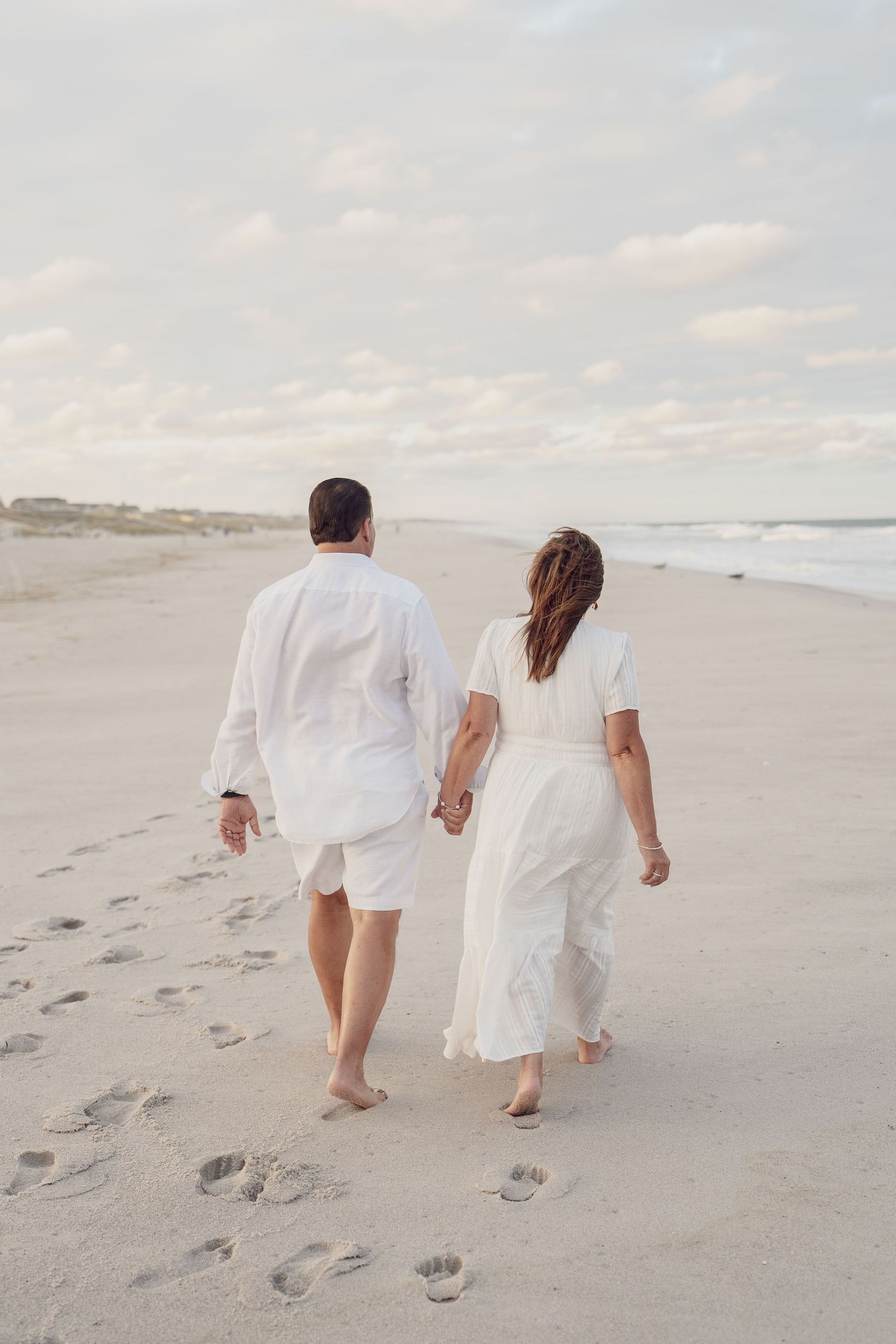 Couple walking barefoot on beach leaving footprints in sand during sunset.