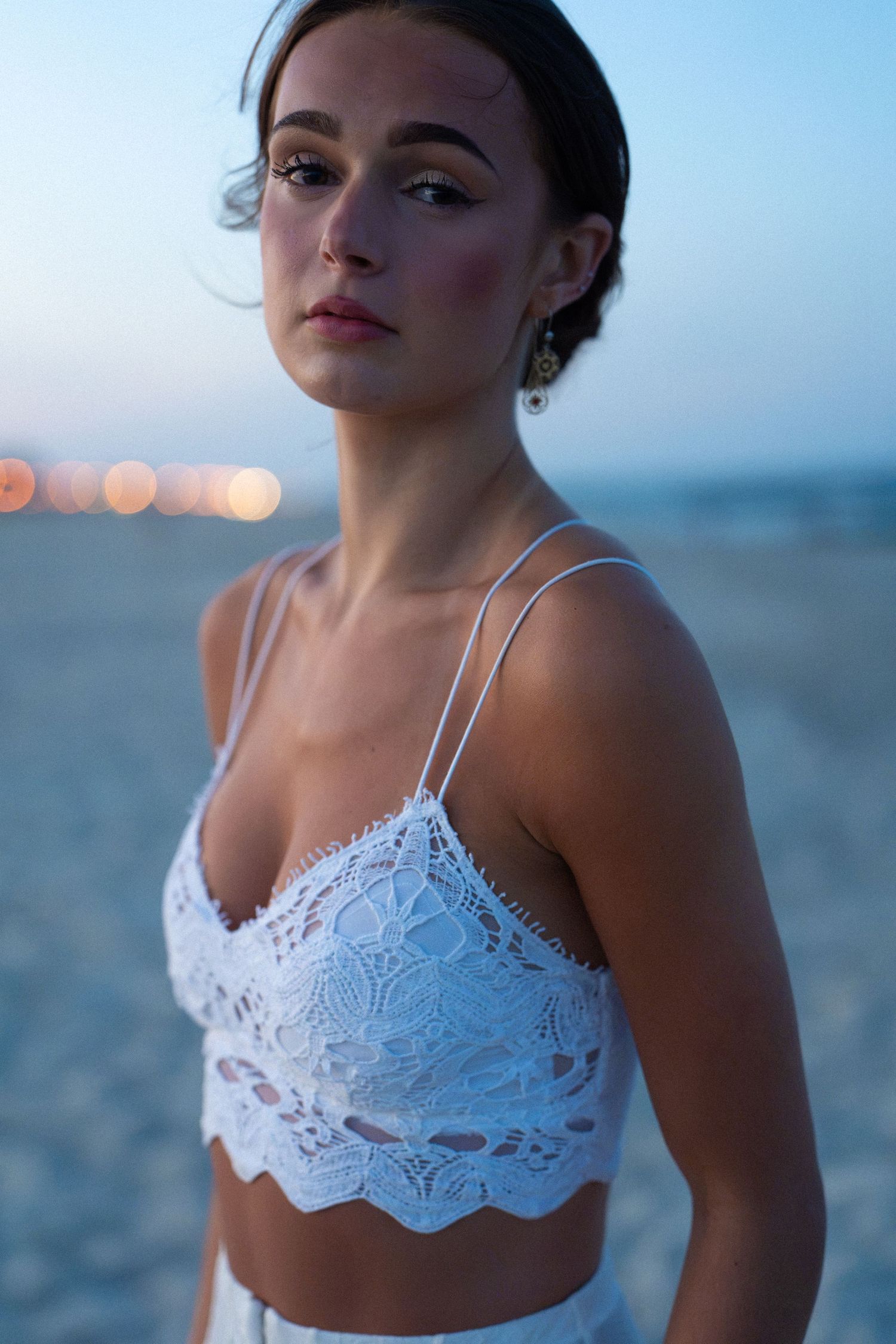 Young woman in white lace bralette top poses on sandy beach at dusk with bokeh lights in background.