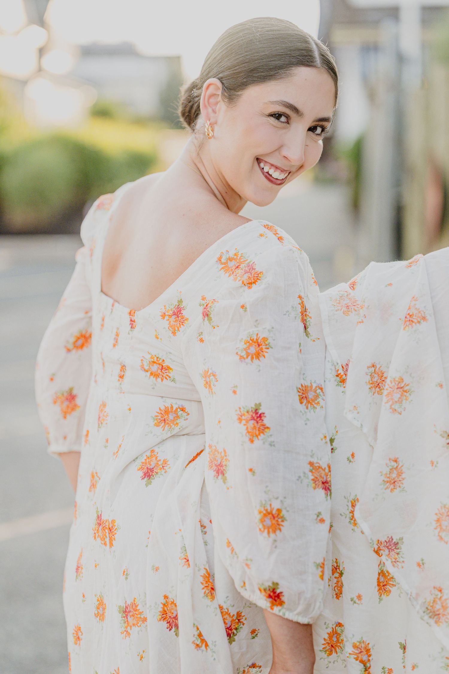 Smiling woman in white floral midi dress with orange blooms, posing outdoors in warm sunlight.