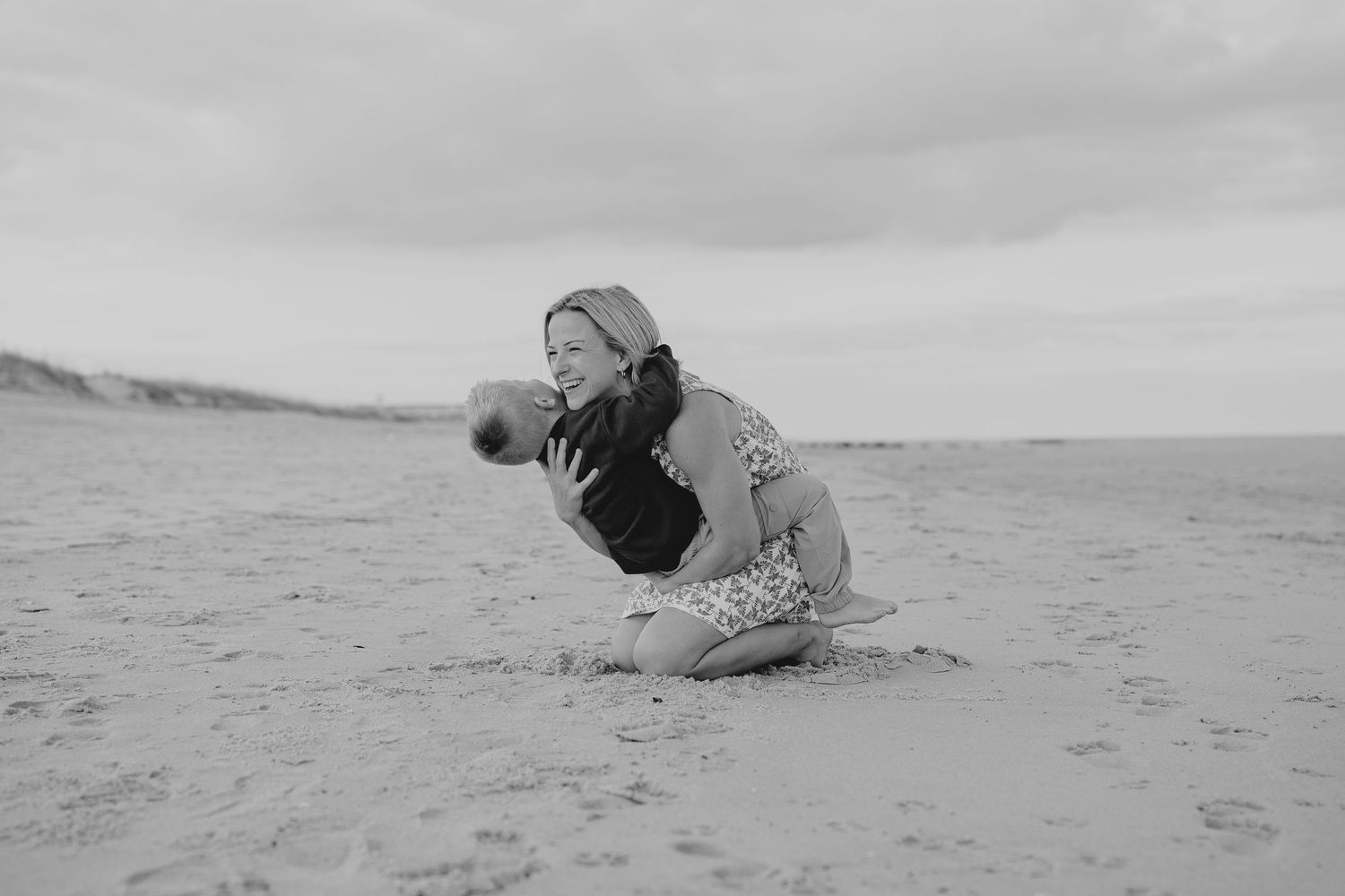 Black and white photo of a woman kneeling on a sandy beach hugging a child on a cloudy day.