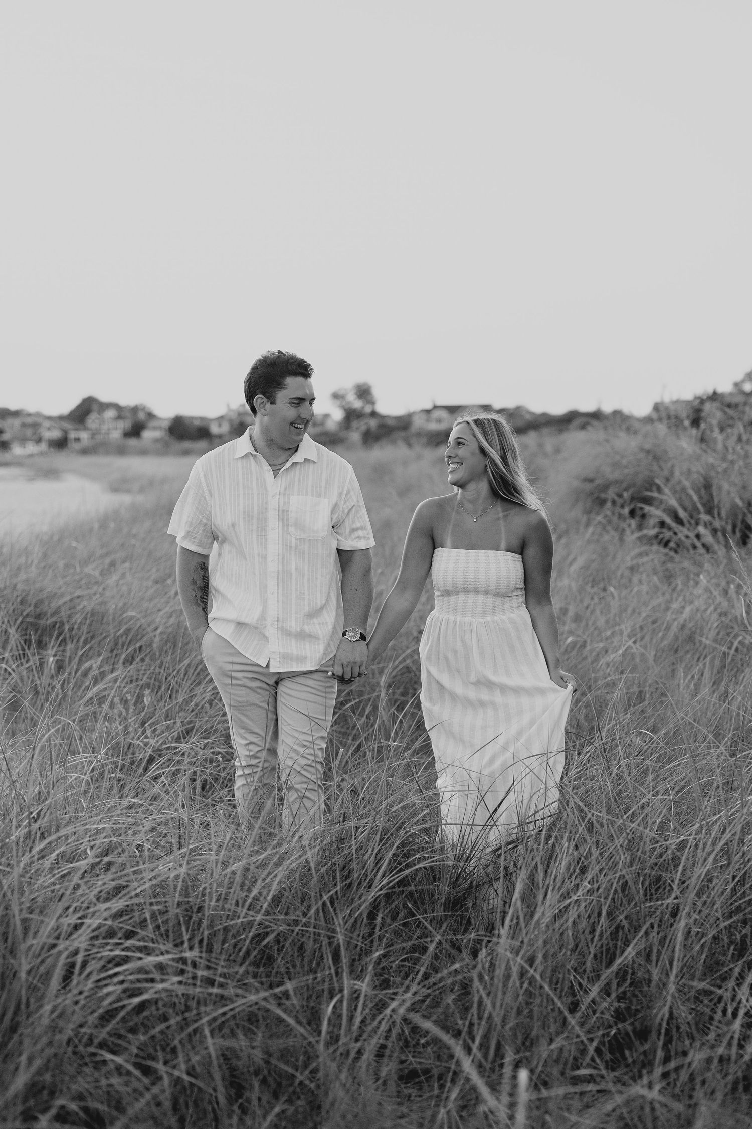 Black and white photo of a couple holding hands and smiling while walking through tall grass near a beach.