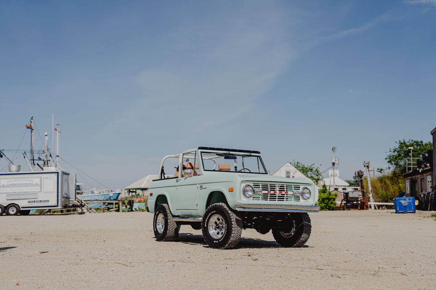 Vintage mint green Ford Bronco parked on sandy ground near a marina under a clear blue sky.