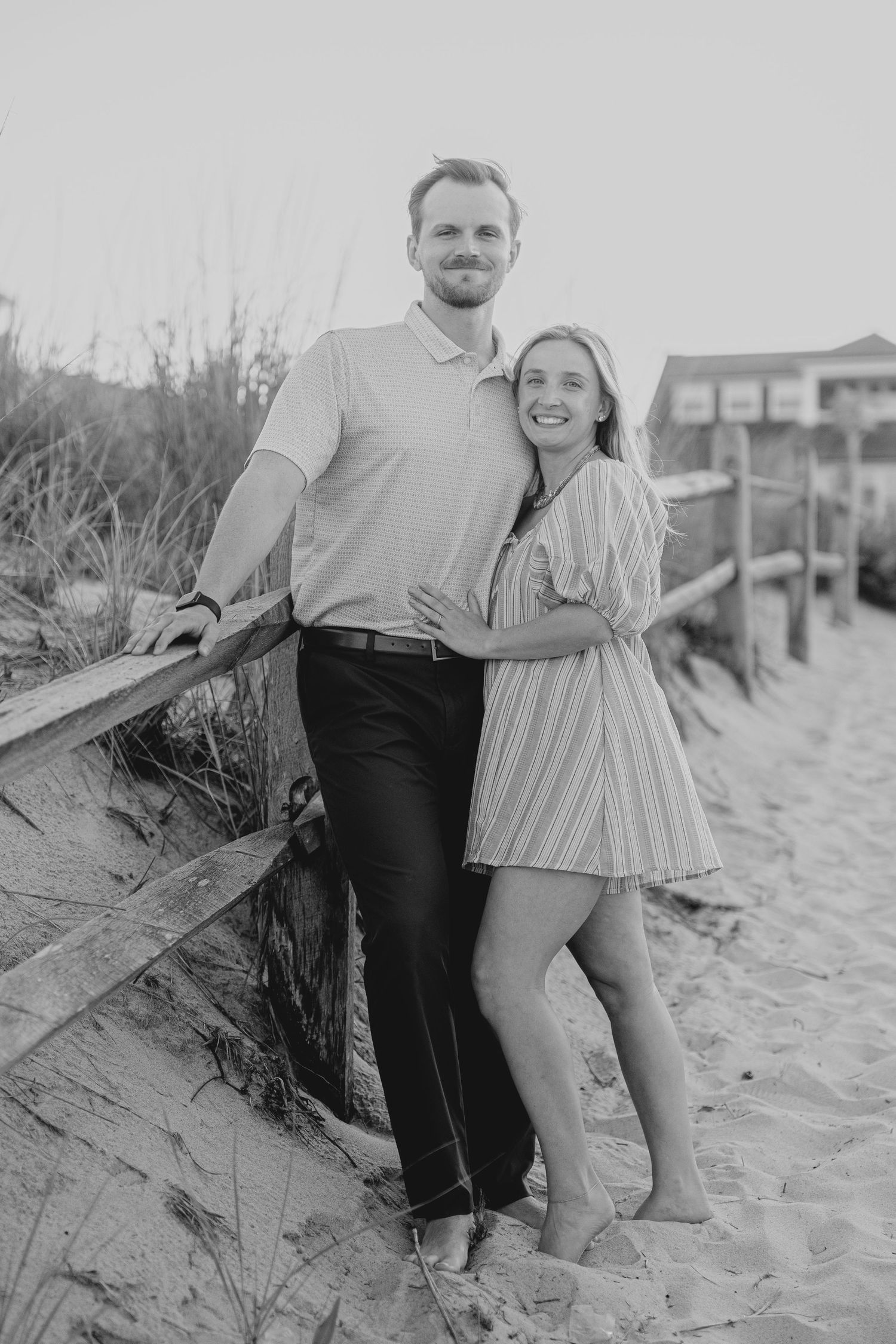 Black and white photo of a smiling couple posing together on a sandy beach path with a wooden fence.