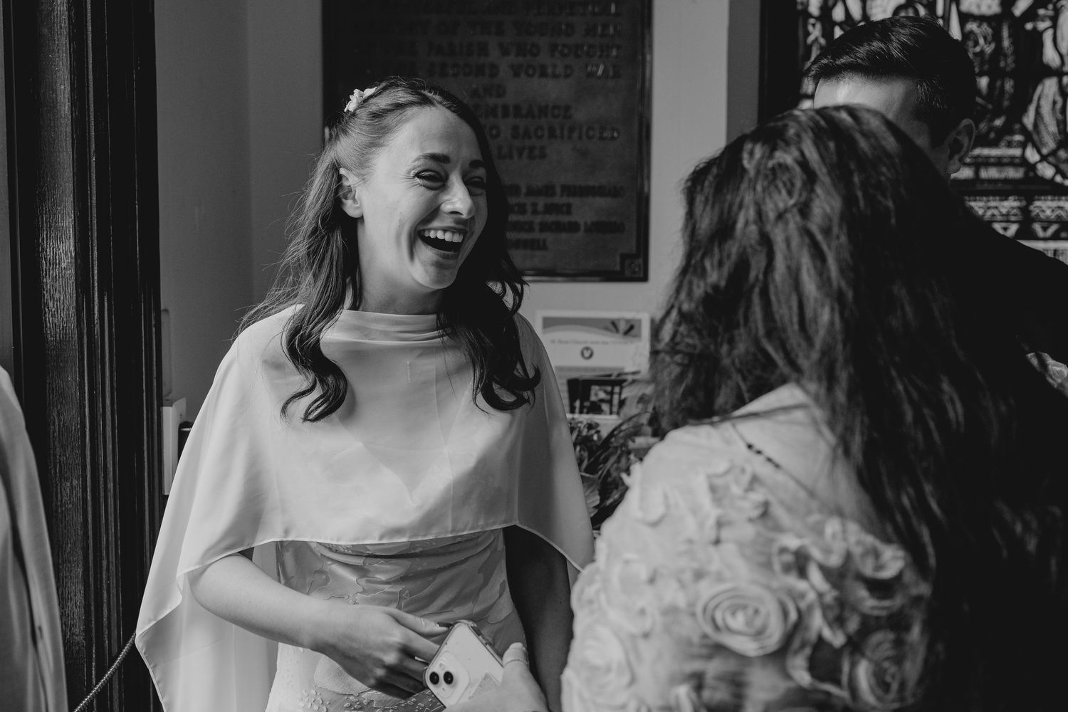 Black and white photo of a laughing bride in elegant gown chatting with a guest at a wedding reception.