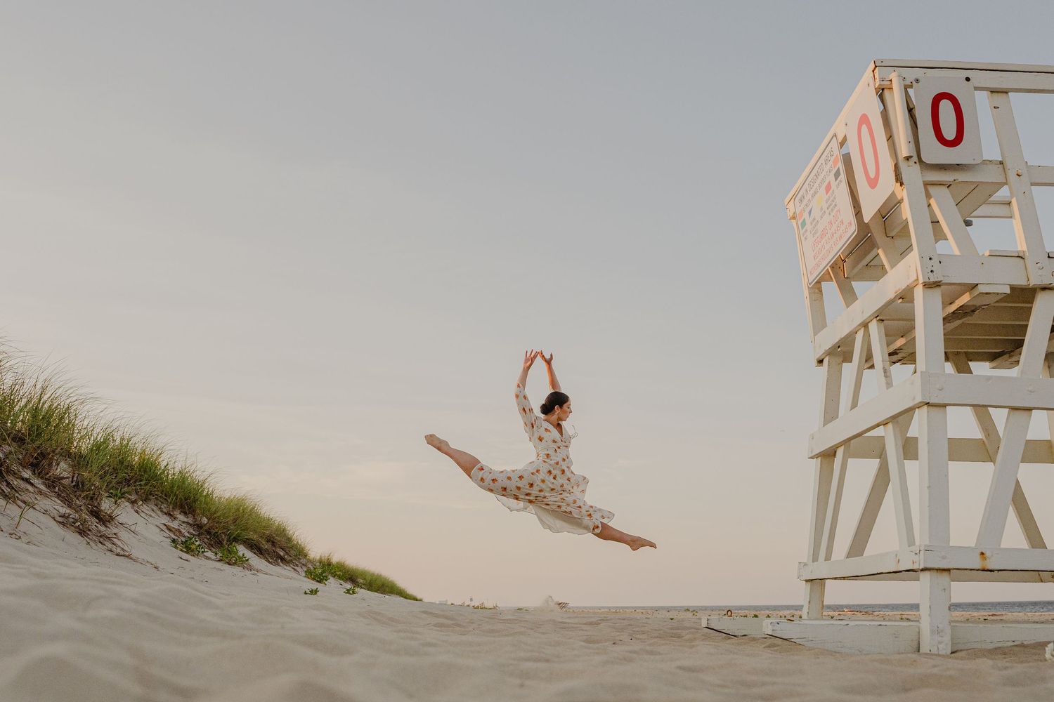 Ballerina performs a grand jeté leap on sandy beach near a lifeguard tower at sunset.