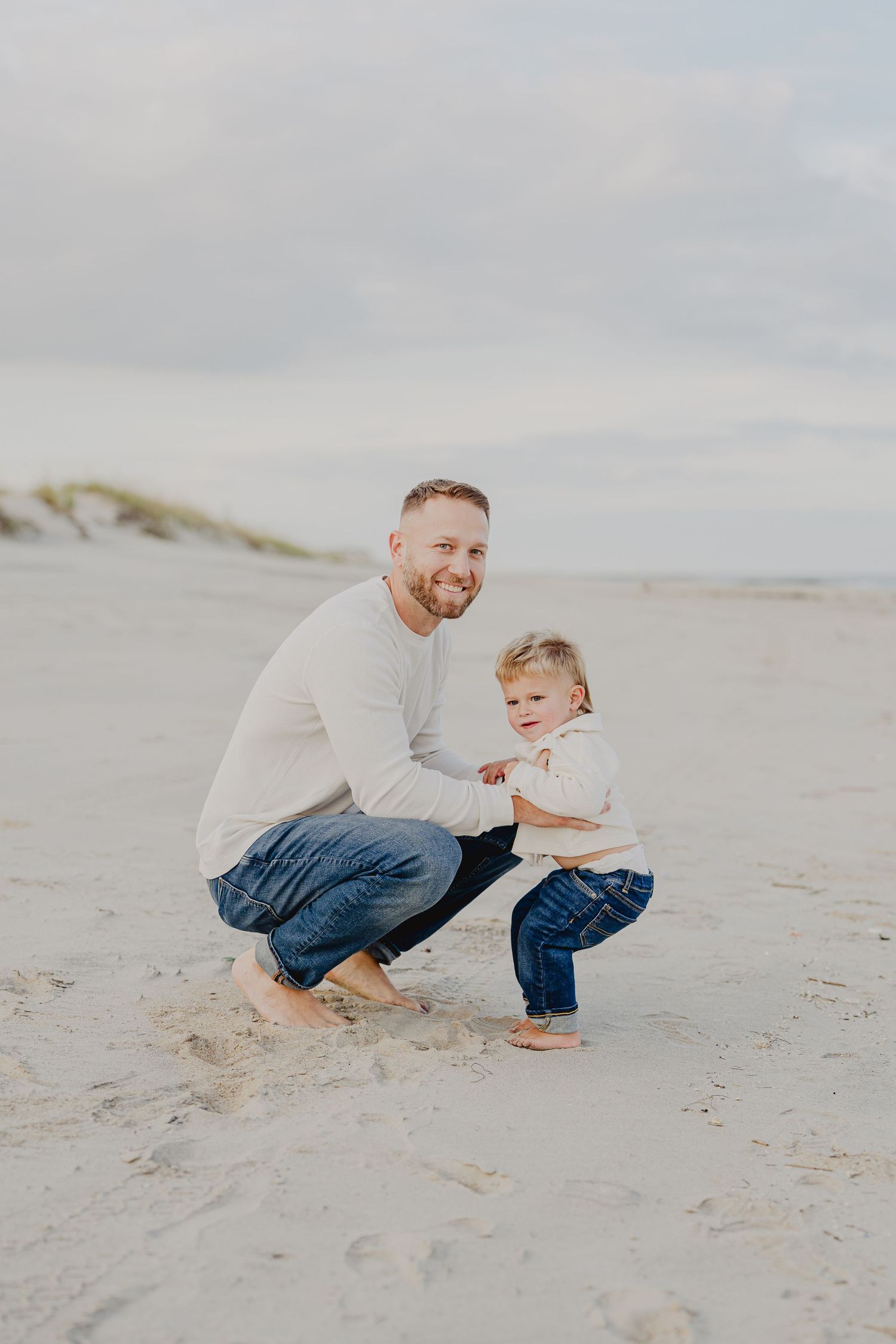 Father and toddler son smiling together barefoot on sandy beach in matching white tops.