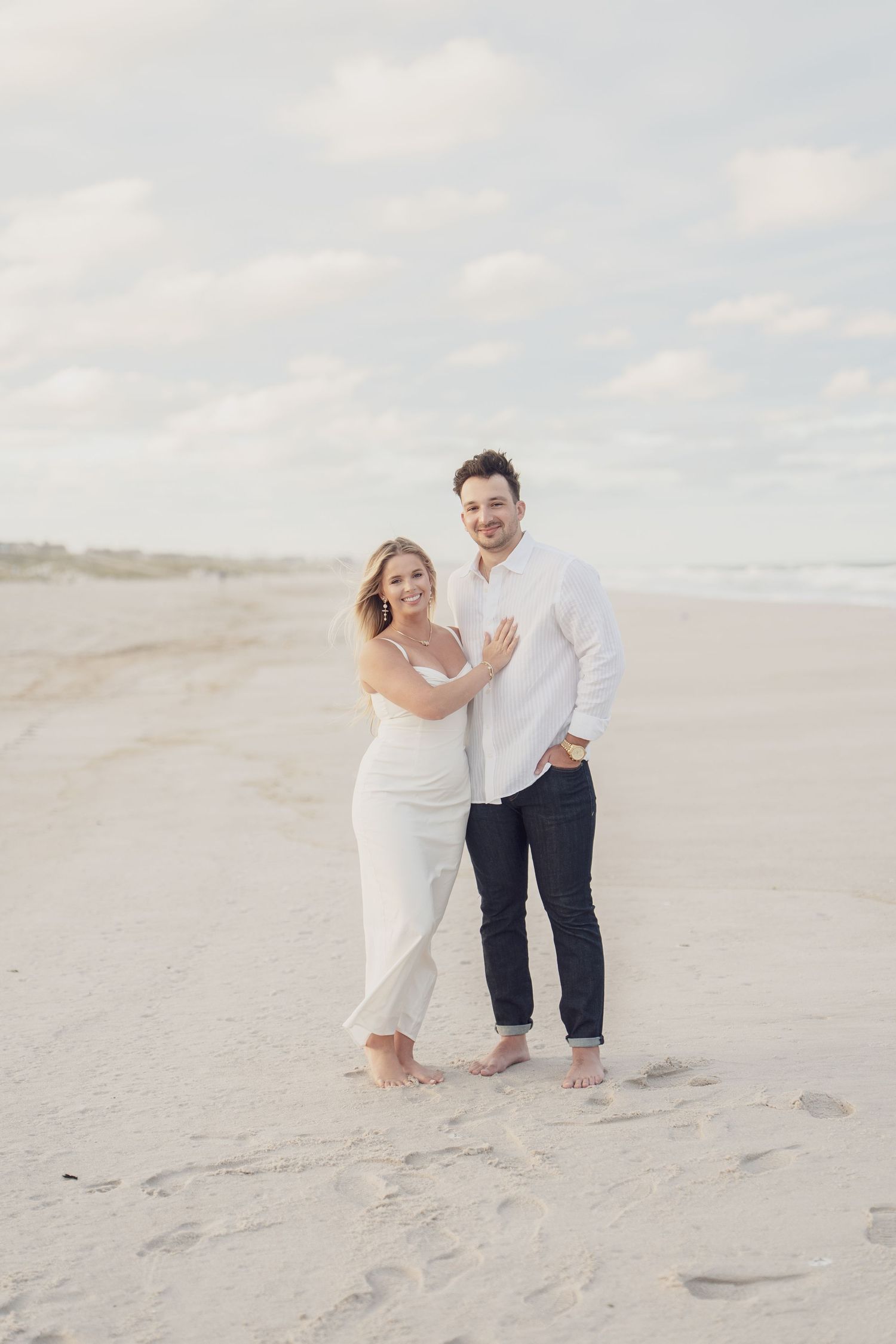 A couple stands barefoot on a peaceful beach at sunset wearing white and navy casual attire.